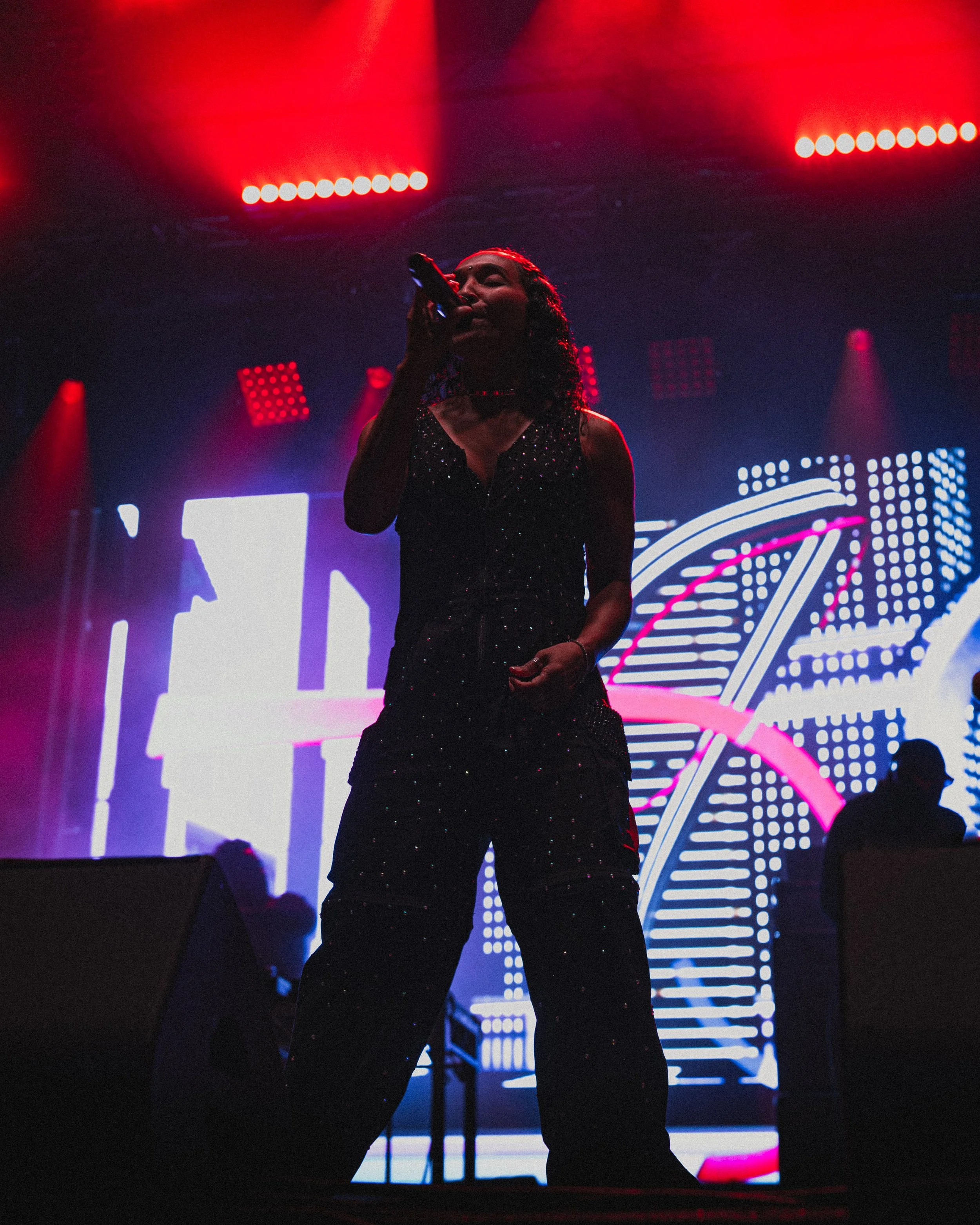 A woman singing on stage with red and blue neon lights and digital graphics in the background.