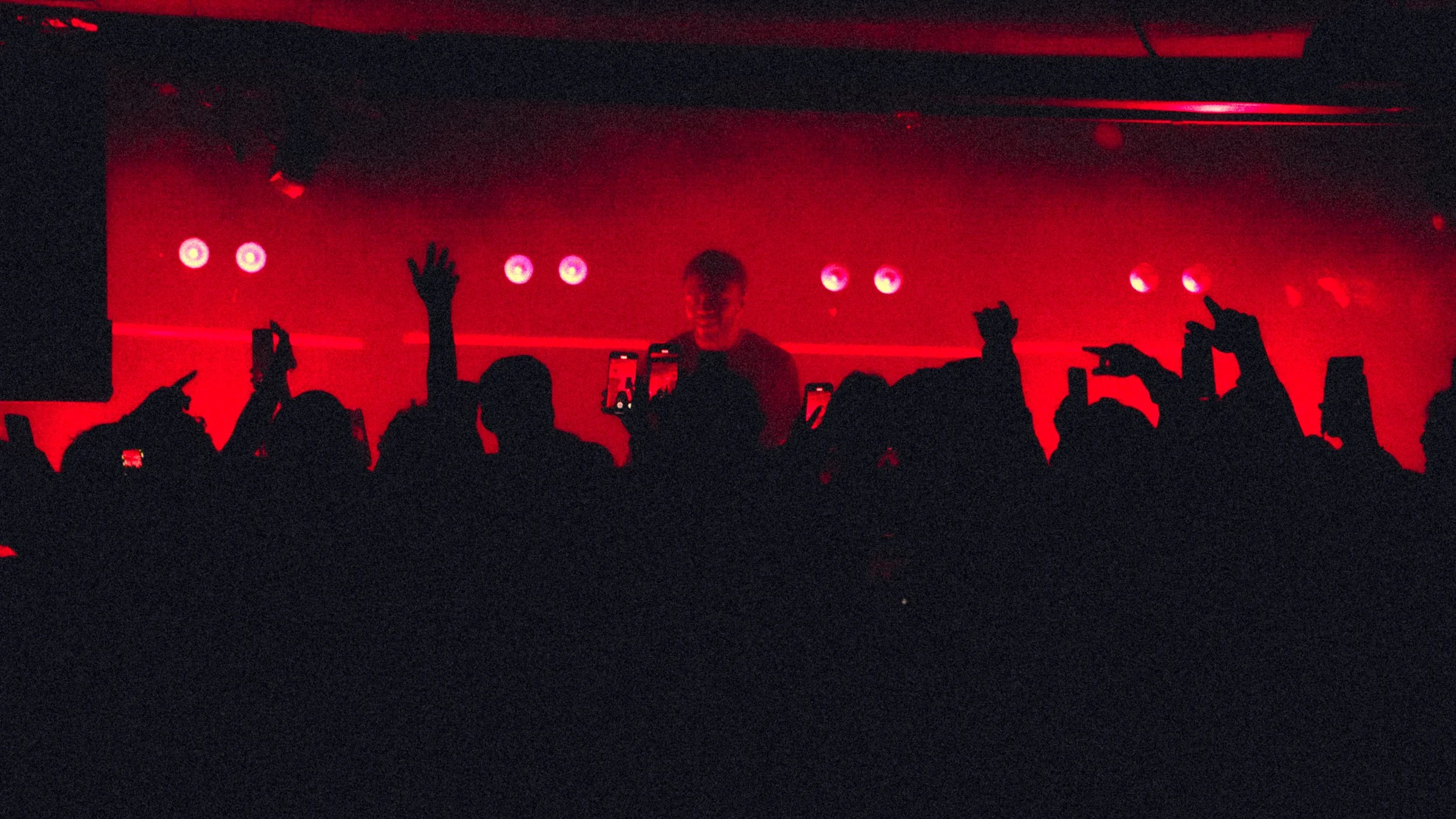 Silhouette of a crowd at a concert with hands raised, facing a performer on stage illuminated with red lights.