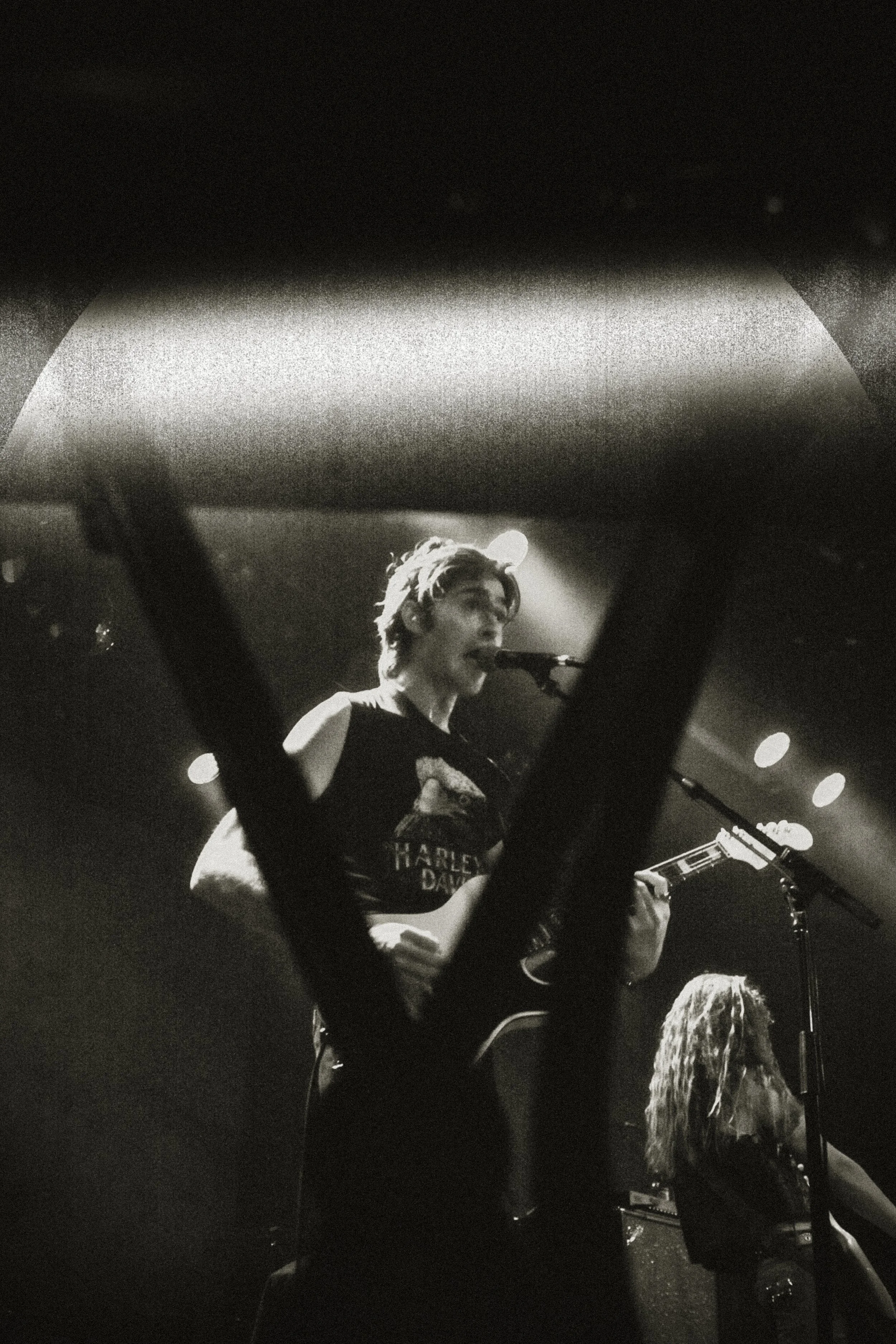 Black and white photo of a female musician singing into a microphone while playing an acoustic guitar on stage. She has short hair and is wearing a Harley Davidson t-shirt. A figure with long hair is visible in the background.