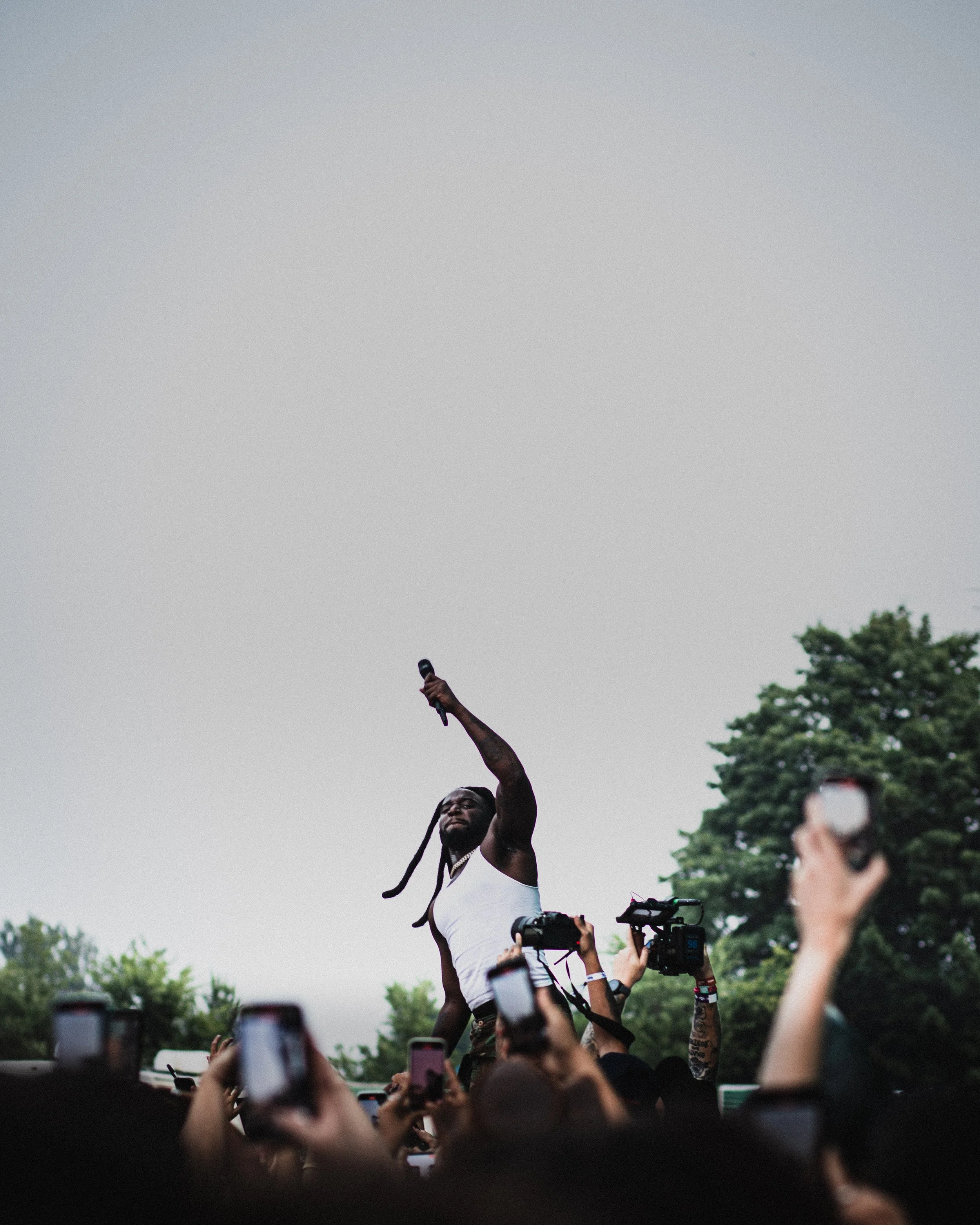 A performer with long braided hair and a tank top holding a microphone raises one arm to the sky during an outdoor concert, surrounded by a crowd taking photos.