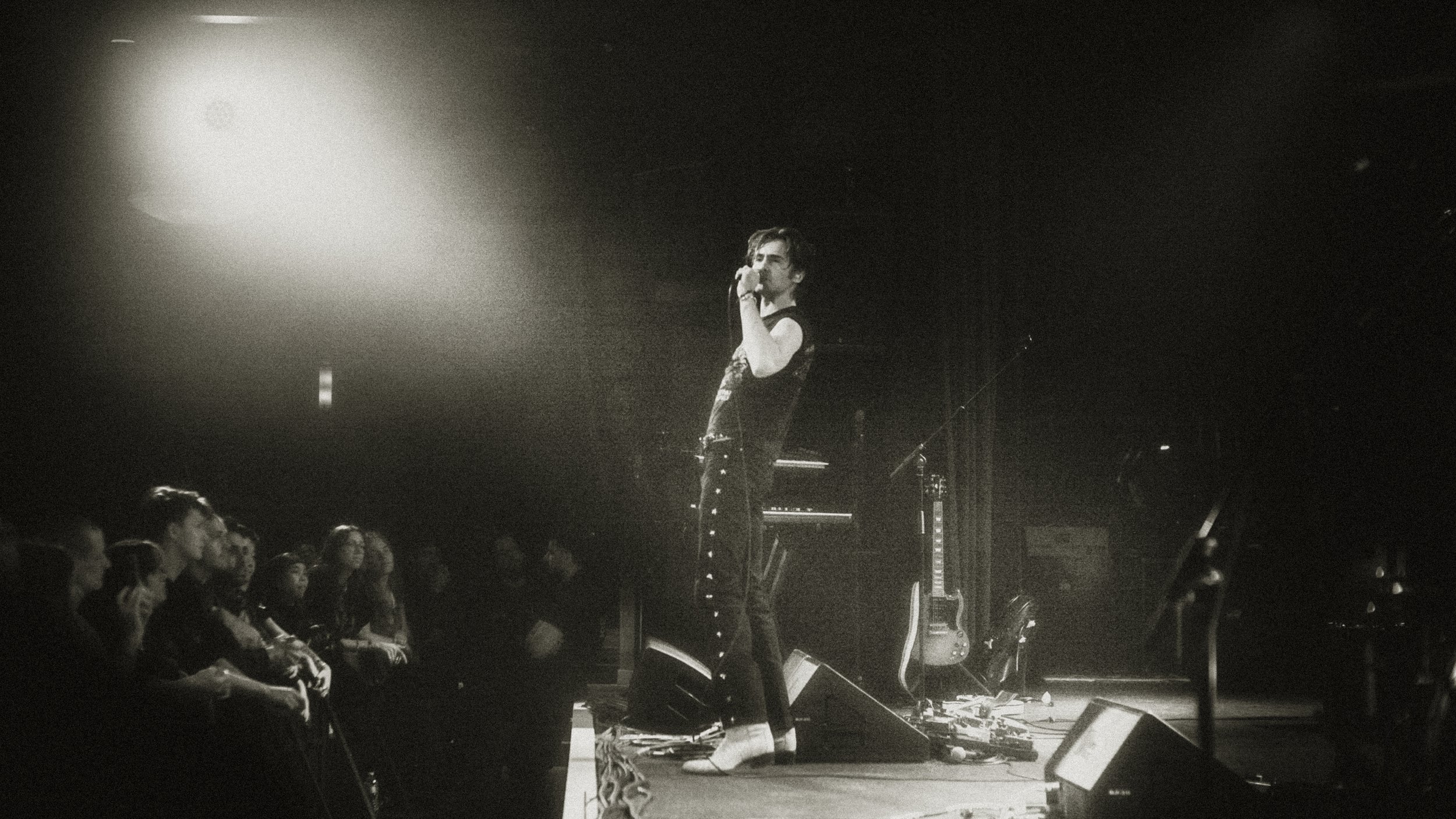 A black and white photo of a singer on stage holding a microphone, wearing a sleeveless top and boots, performing in front of an audience. There is music equipment, including guitars and a keyboard, visible behind the singer.