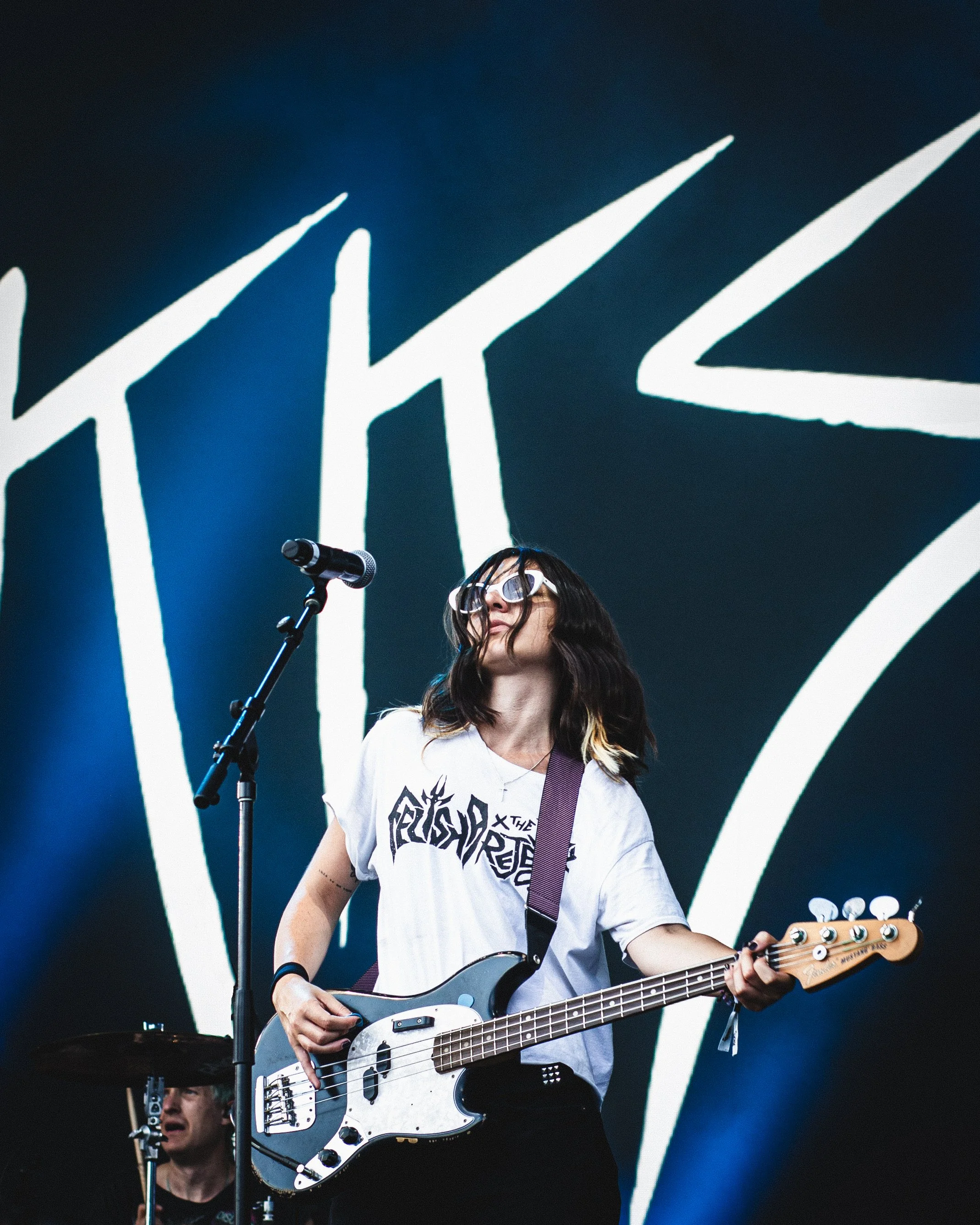 Female guitarist playing bass guitar on stage with a large band logo in the background.