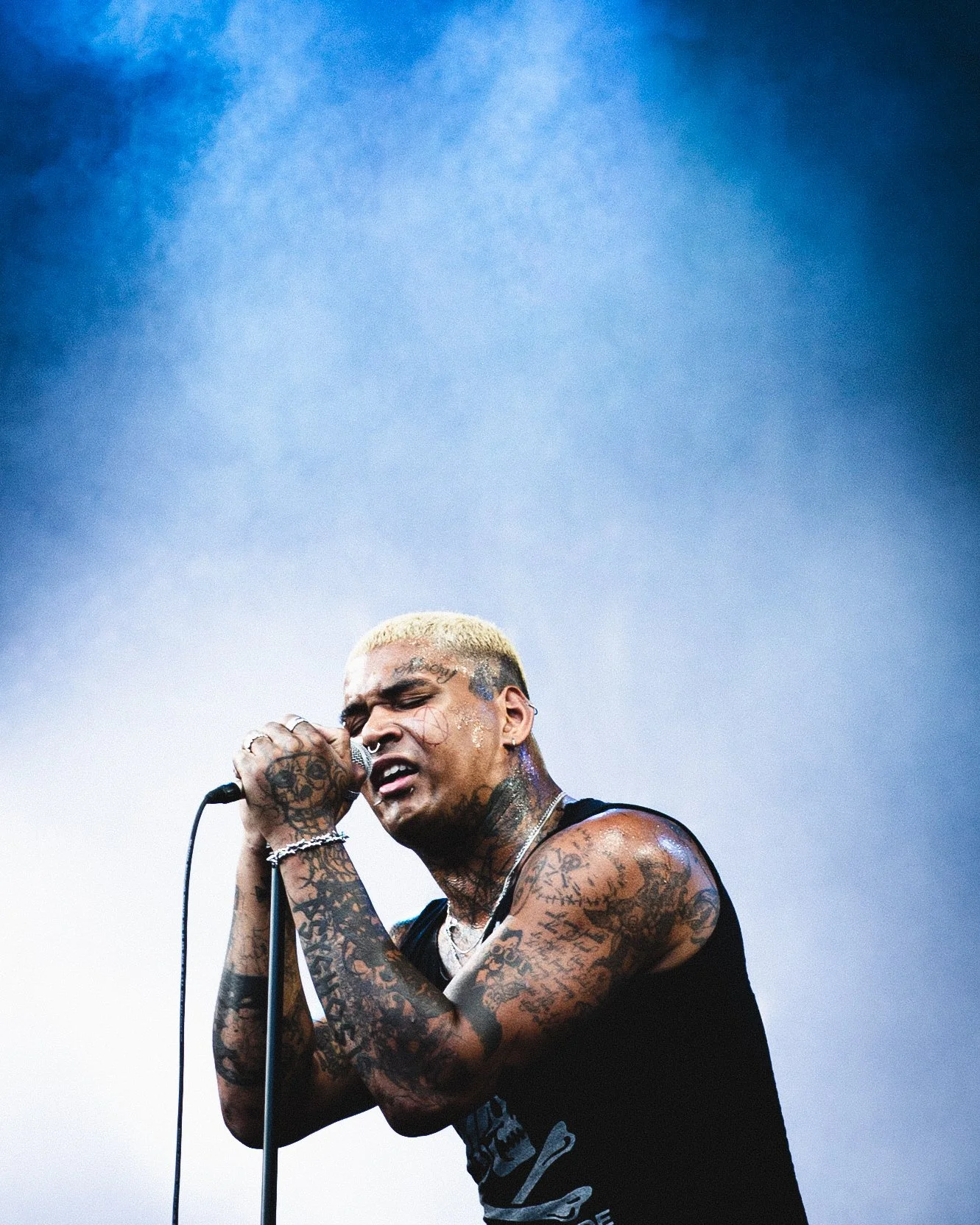 A male singer with tattoos on his arms and face, holding a microphone and performing passionately on stage, with a blue background.