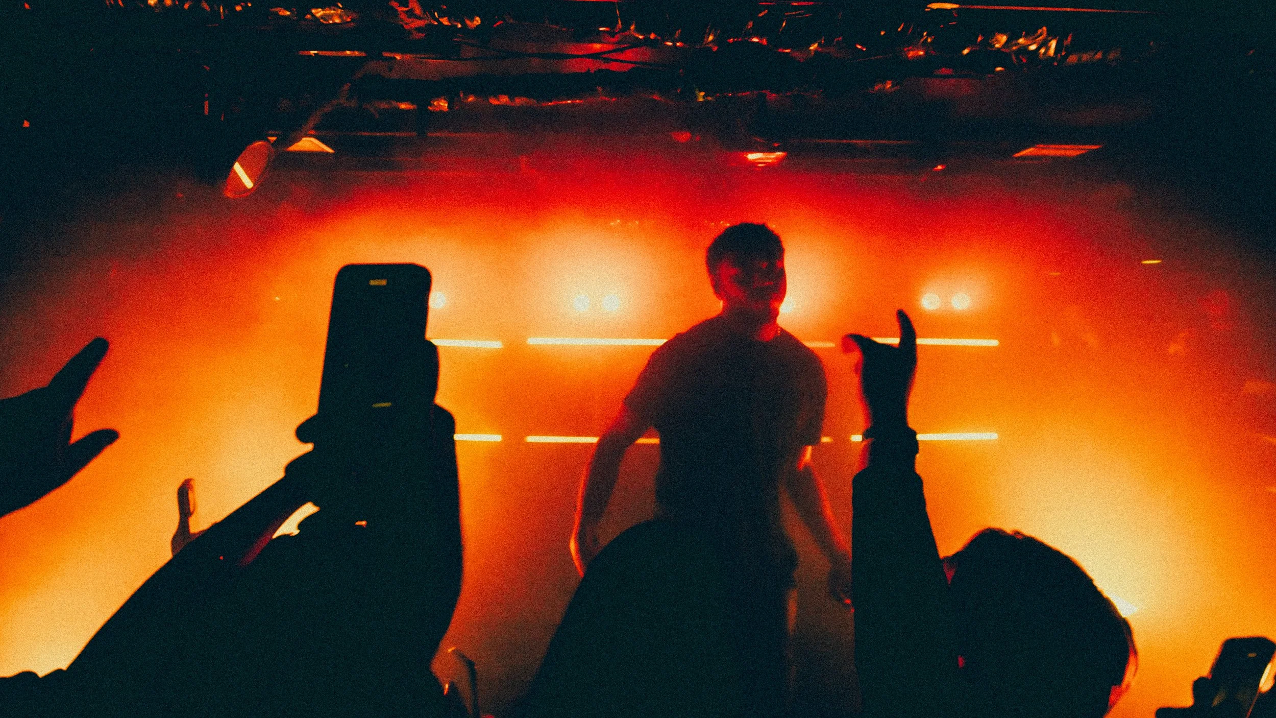 A performer on stage in front of orange stage lighting, with audience members raising their hands and taking photos.