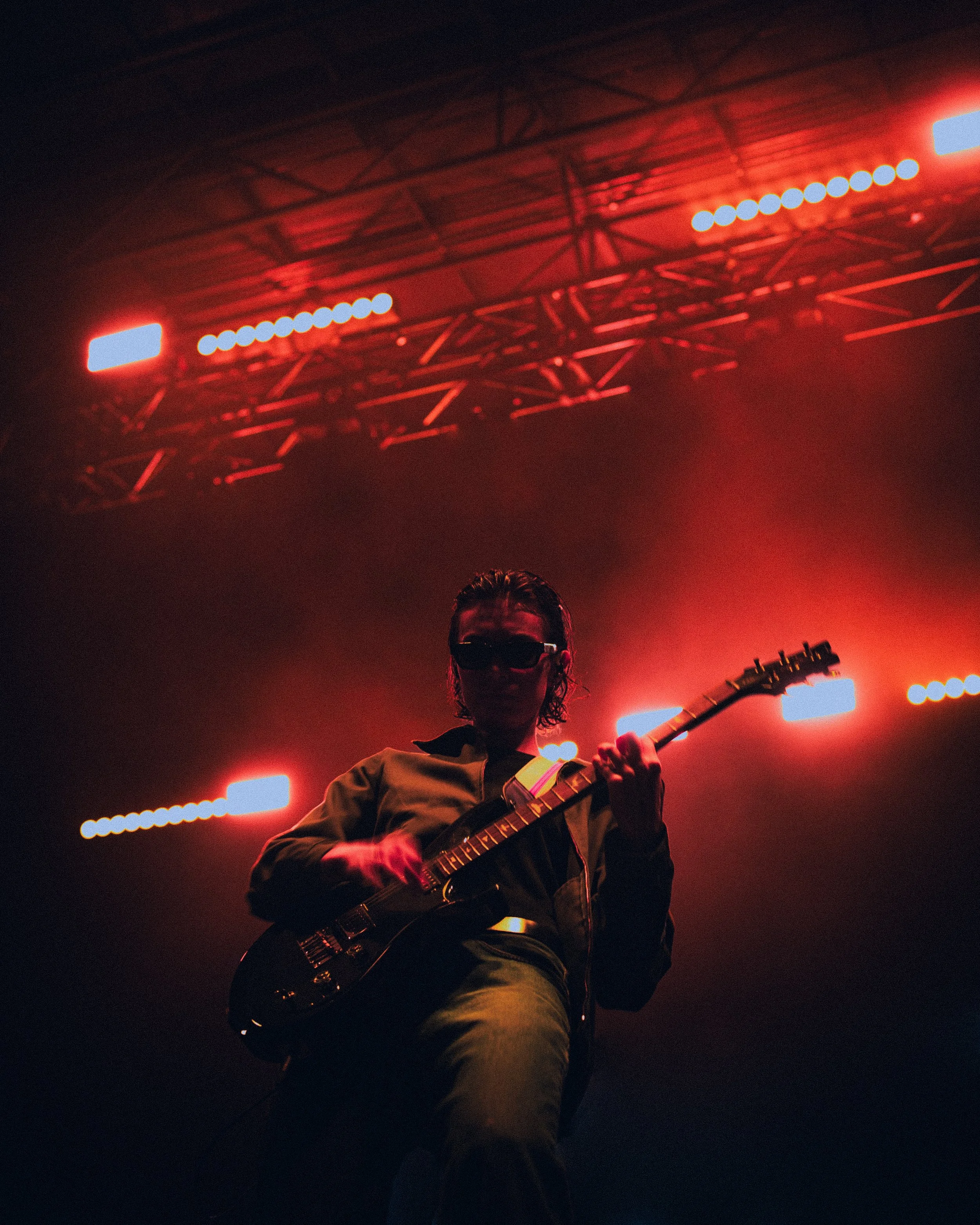 A musician playing an electric guitar on stage under red lights with a dark background.