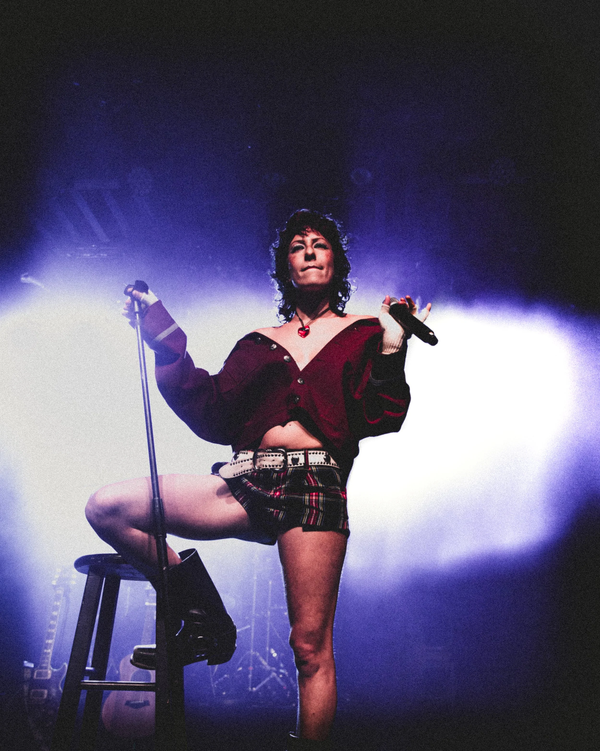 A woman standing on stage with one leg raised on a stool, holding a microphone and a wireless microphone, with purple and white stage lights behind her.