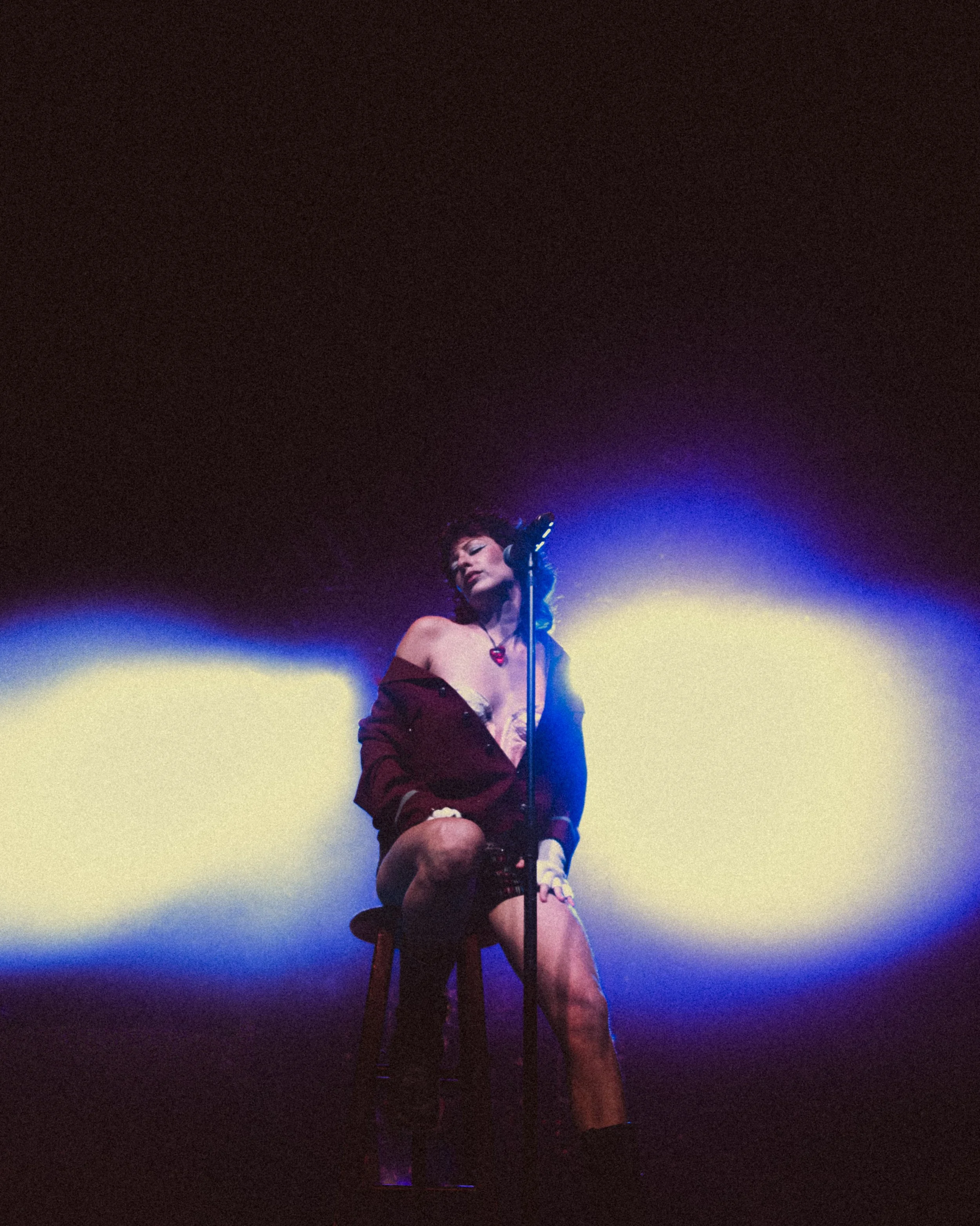 A woman sitting on a stool singing into a microphone with colorful stage lights shining behind her.