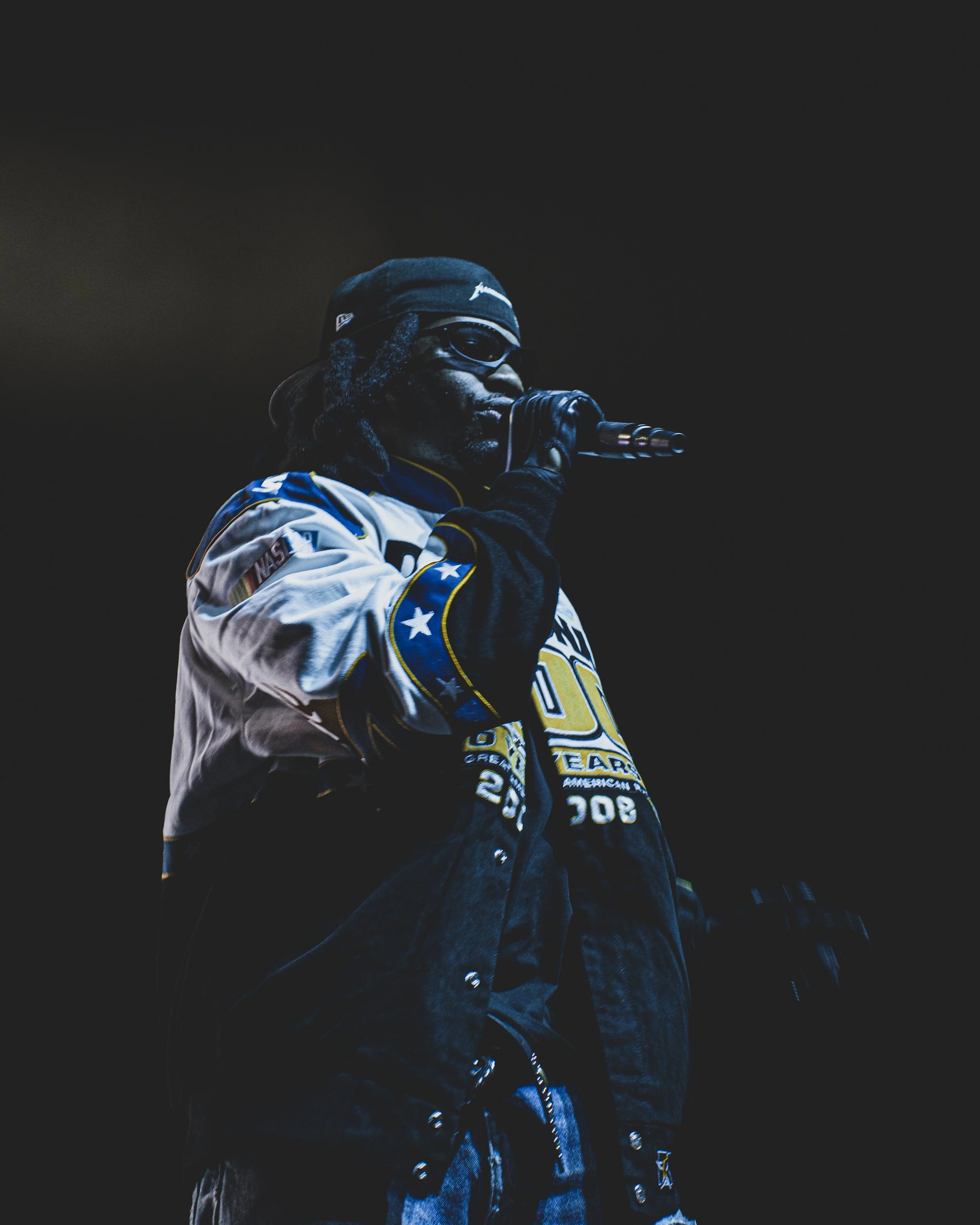 A man with dreadlocks, wearing sunglasses, a camouflage cap, and a hockey jersey, is holding a microphone and performing on stage against a dark background.