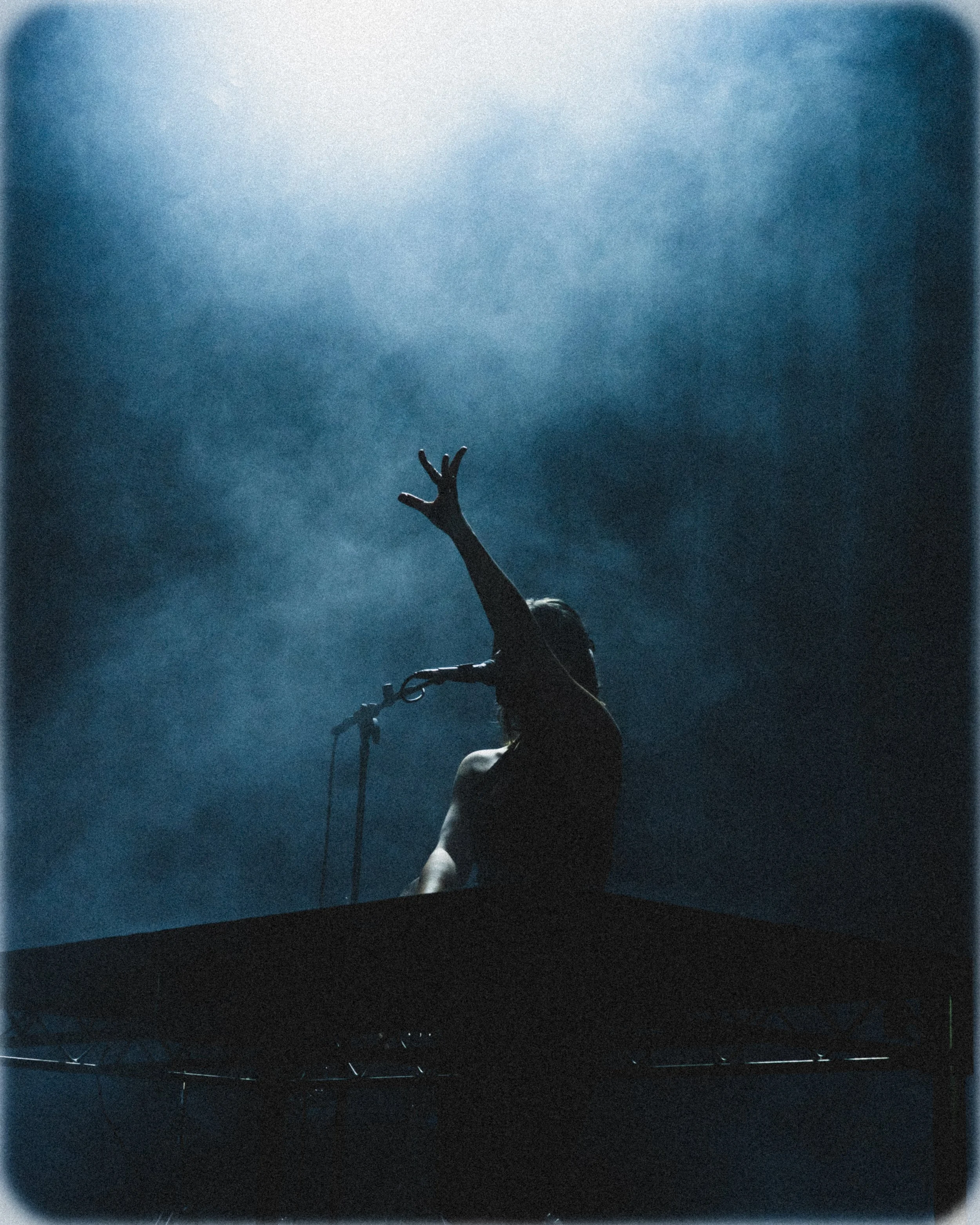 Silhouette of a person with long hair, standing behind a music keyboard, raising one arm with fingers extended, against a blue-tinted smoky background.