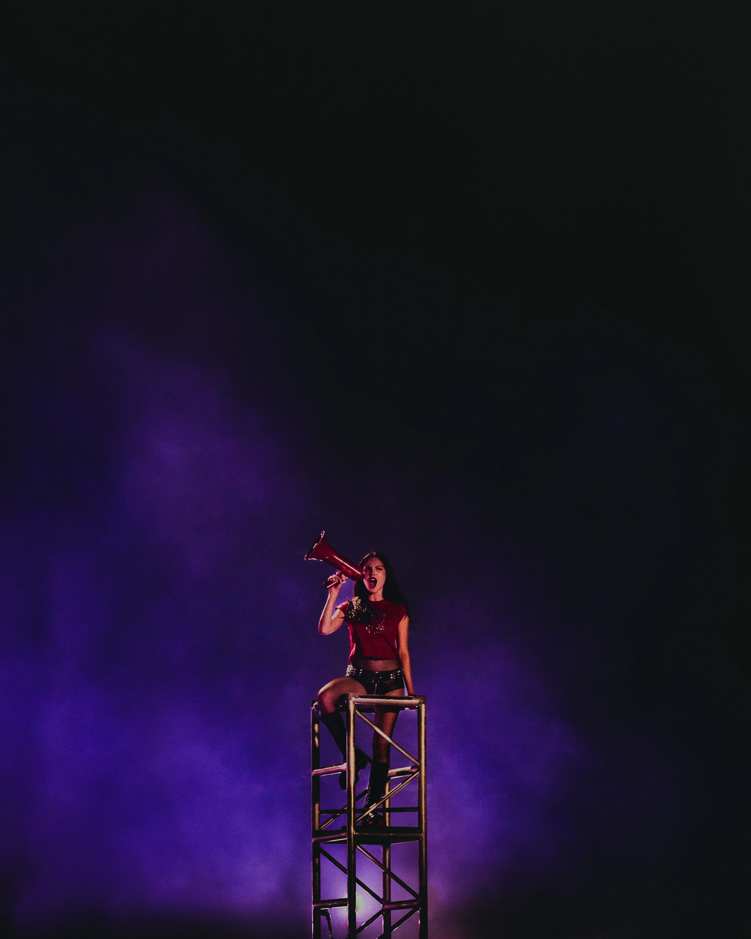A woman on a tall metal ladder holding a red megaphone to her mouth, singing or shouting against a dark background with purple lighting and smoke.
