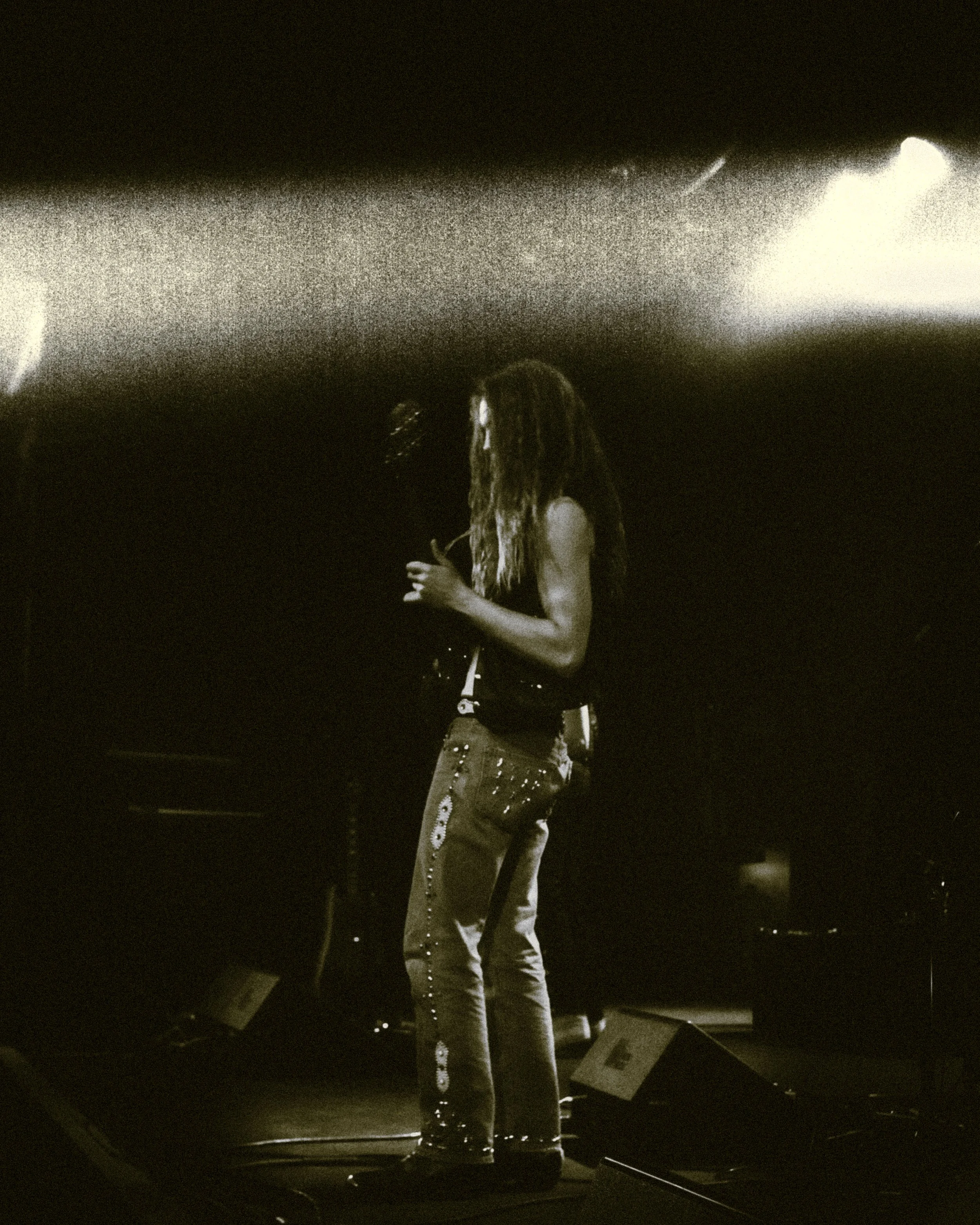 A person with long hair standing on a stage, looking at a phone, with stage equipment and monitors around.