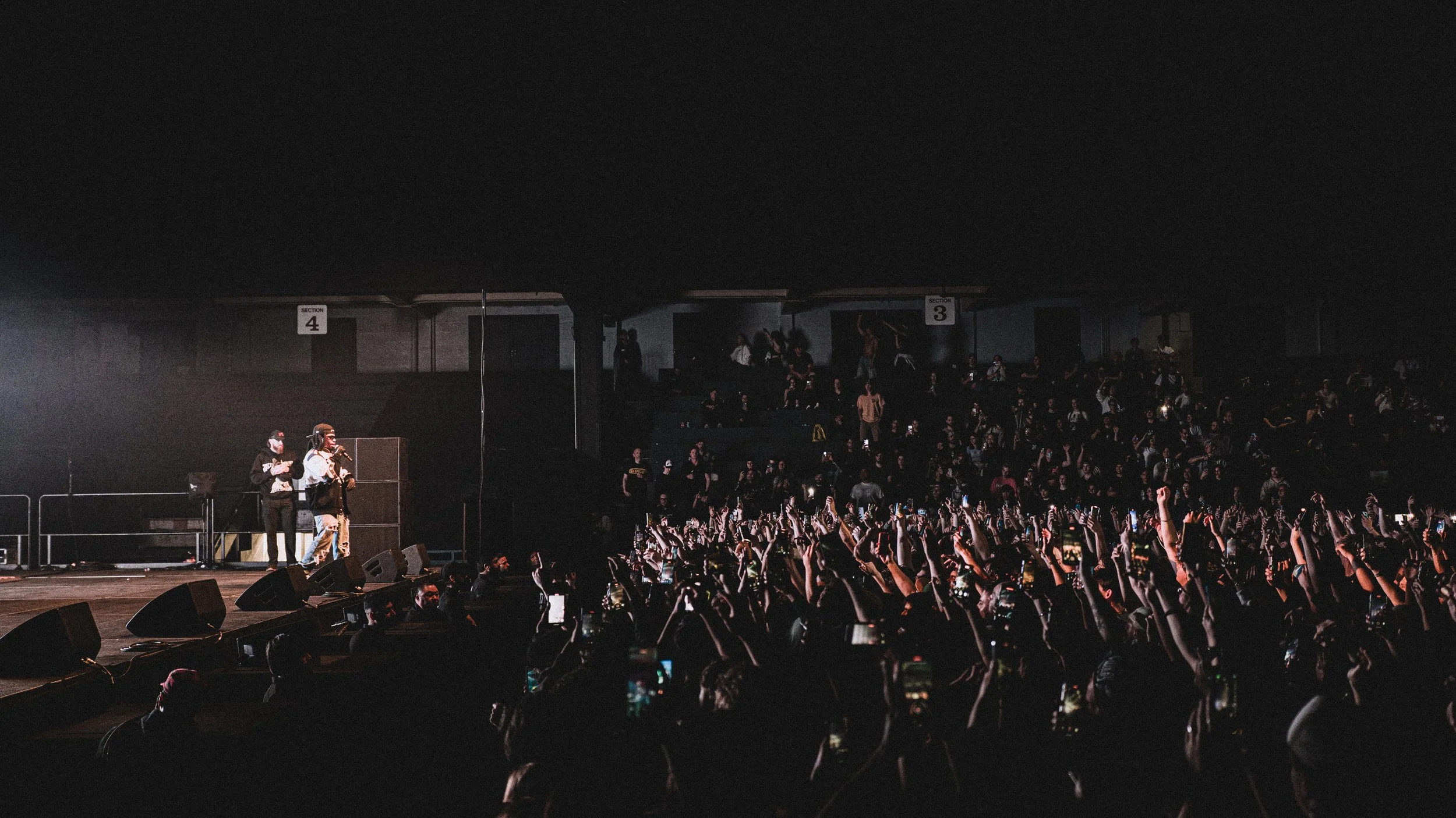 A concert scene with a crowd of fans raising their hands and holding up smartphones, facing a stage with performers, in a dark indoor venue.