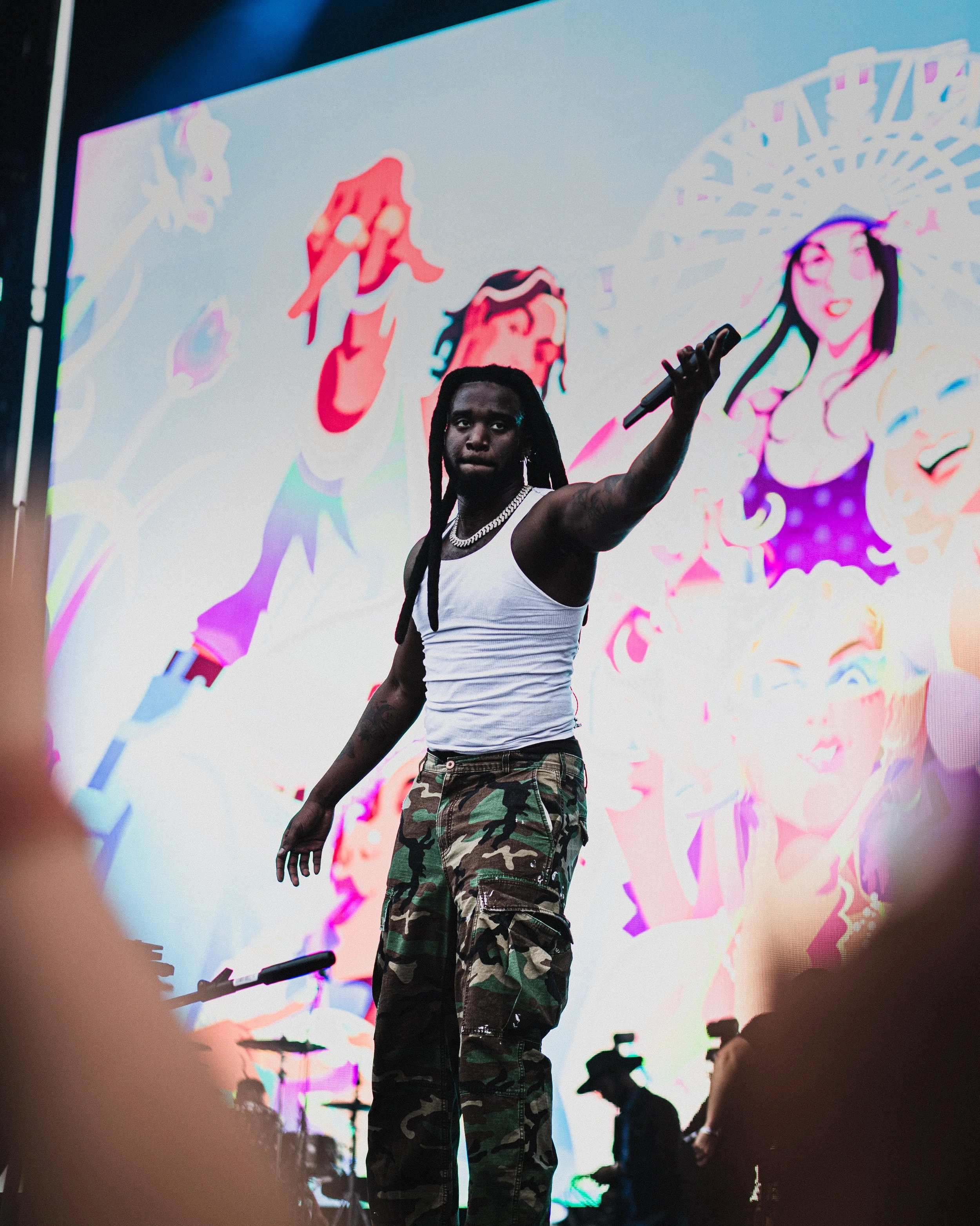 A male performer with long dreadlocks, wearing a white tank top, camouflage pants, and a silver chain, holding a microphone on stage with a digital screen in the background displaying colorful cartoon-like characters.