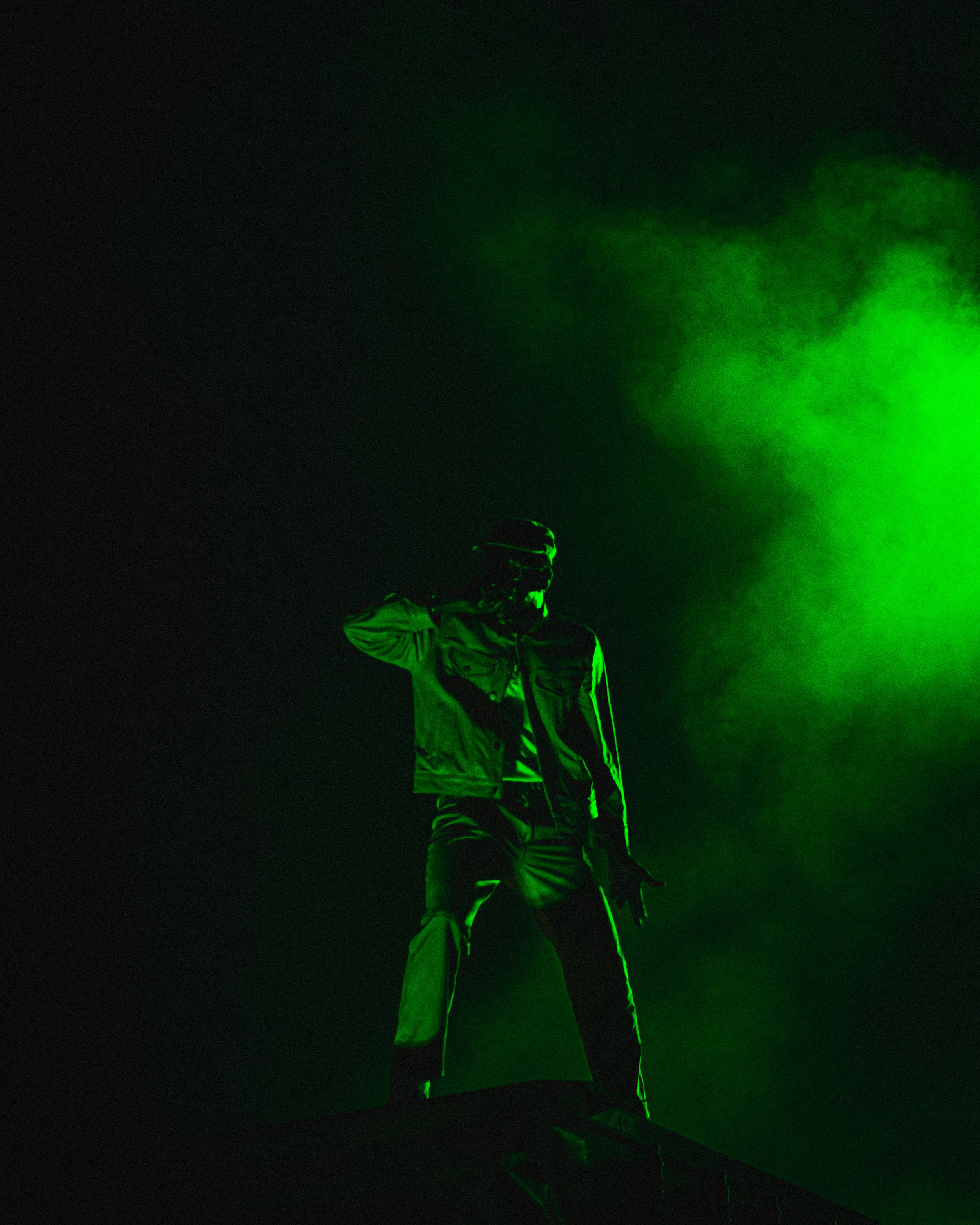Person wearing sunglasses and a jacket, standing on a dark stage illuminated by green light and smoke.