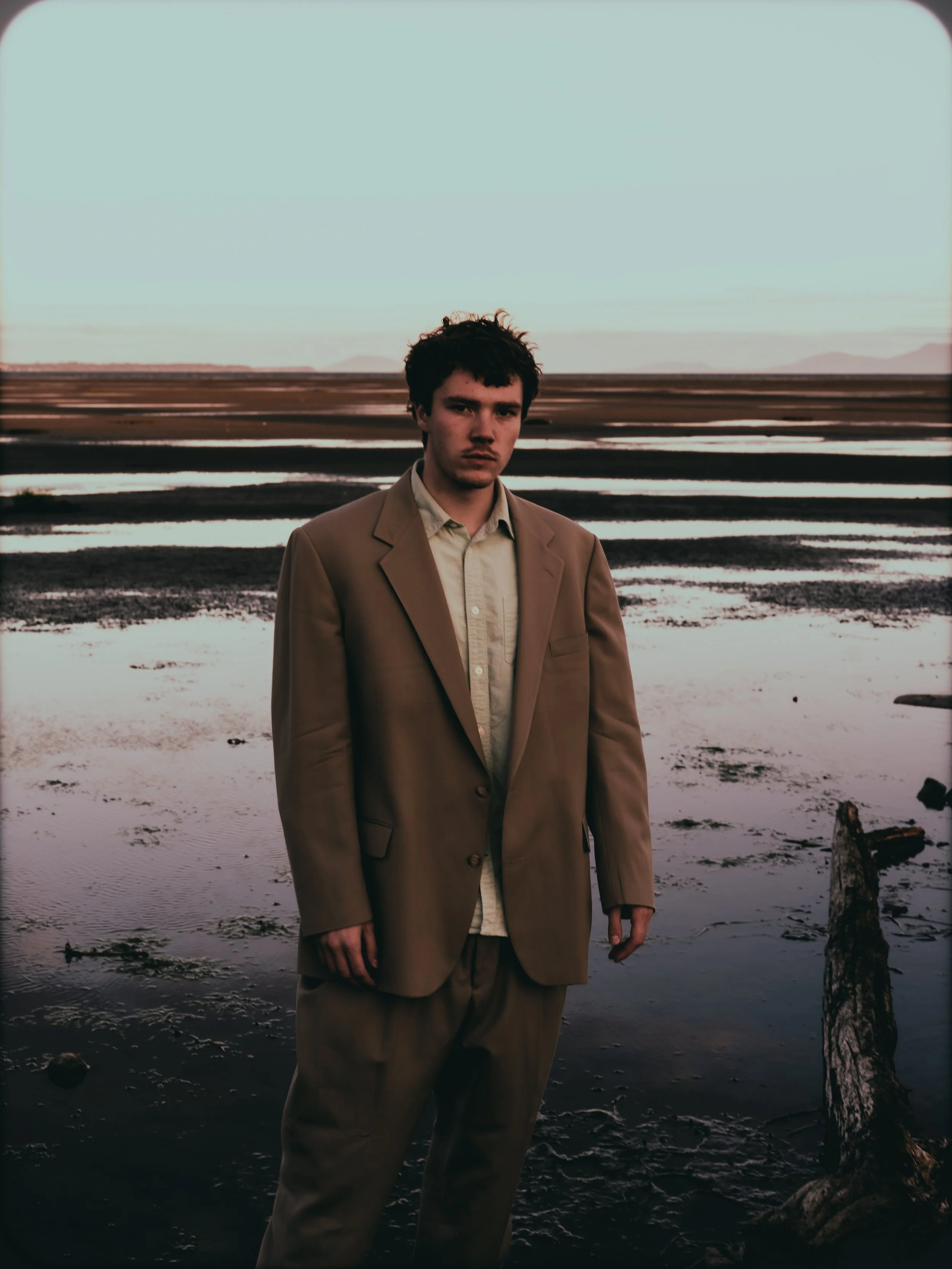 A man wearing a brown suit standing on a wet, muddy shoreline at sunset with a desolate landscape and distant mountains in the background.