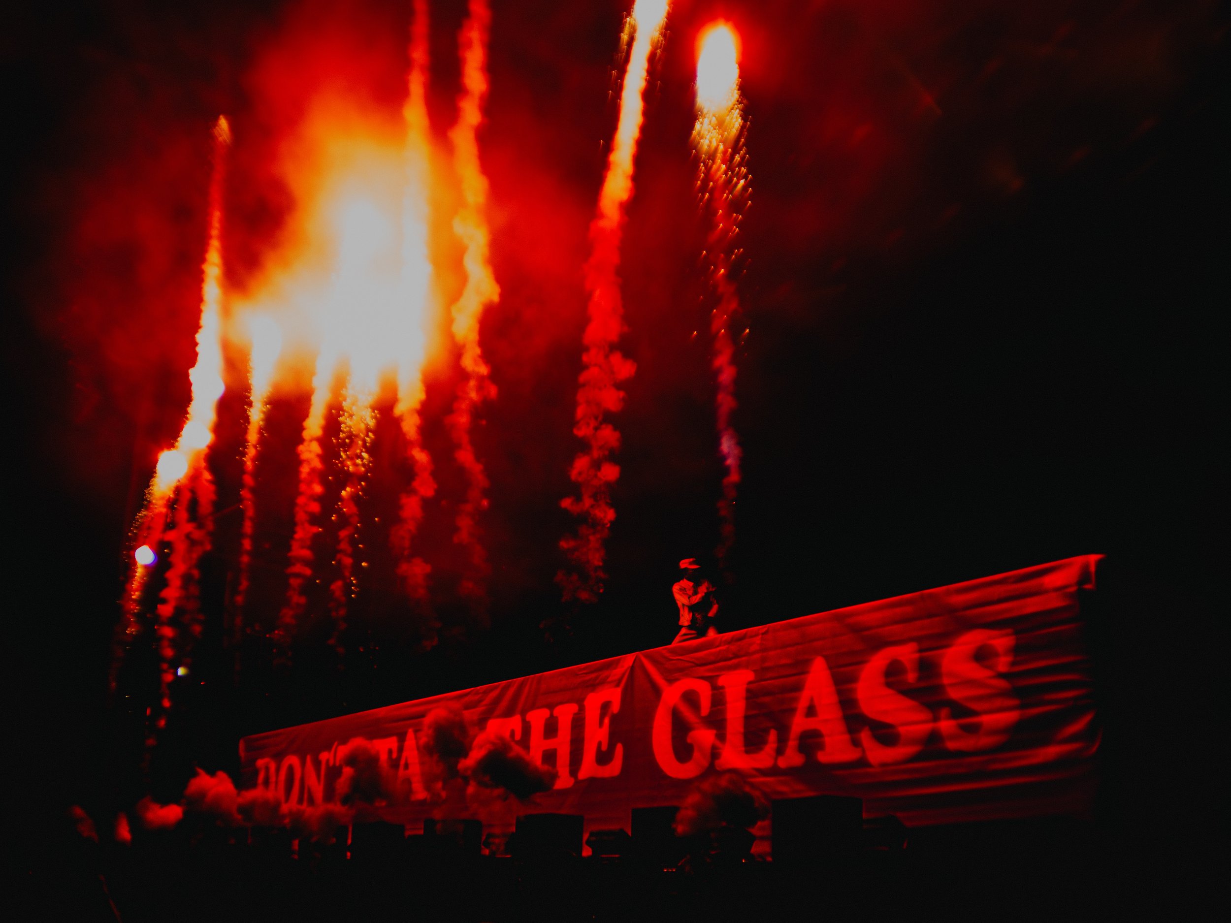 Fireworks lighting up the night sky above a protest banner that reads 'DON'T TREAT THE GLASS' with a person standing on top of the banner.