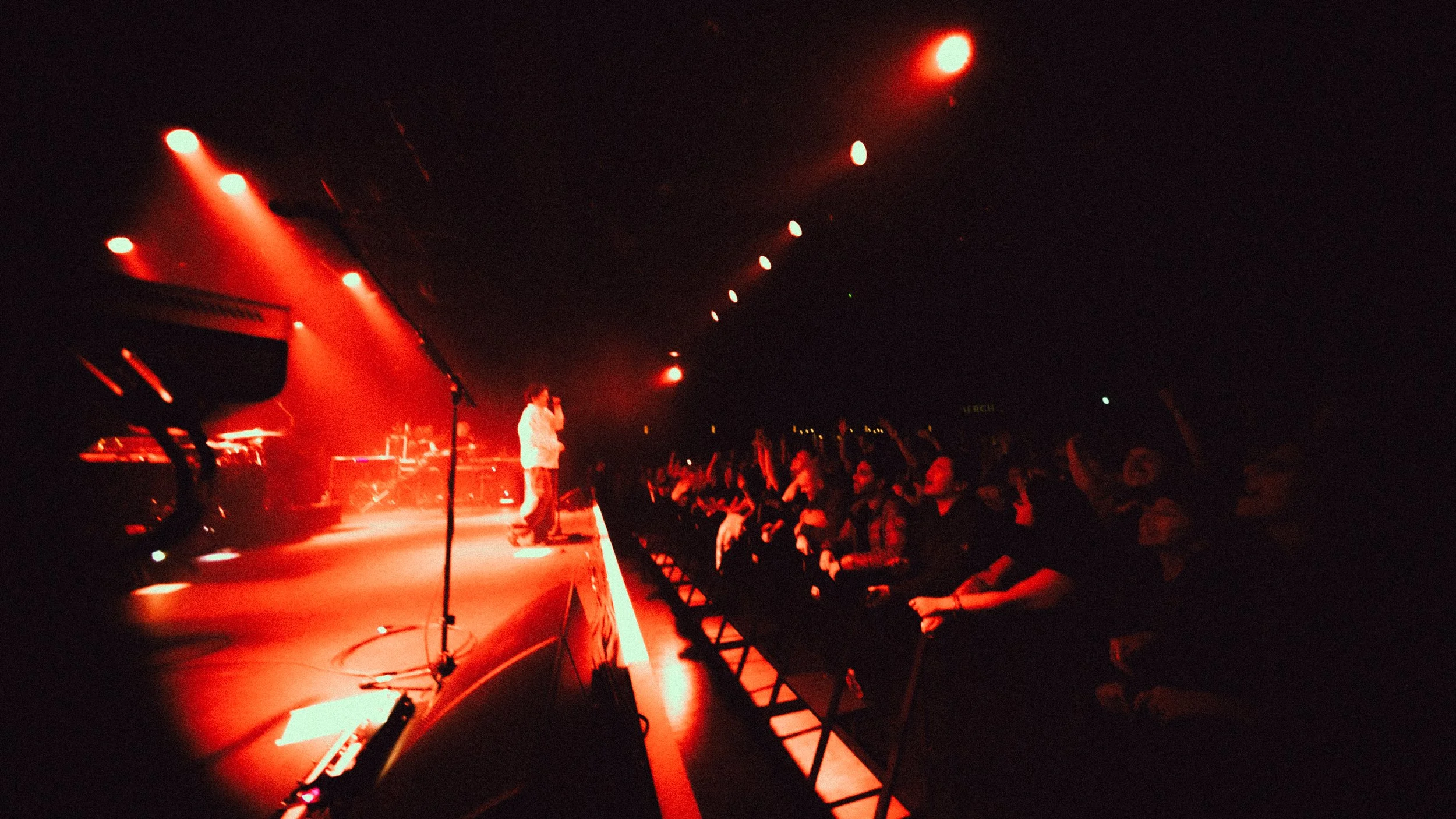 Concert scene with a performer on stage under red lights and an audience raising their hands.