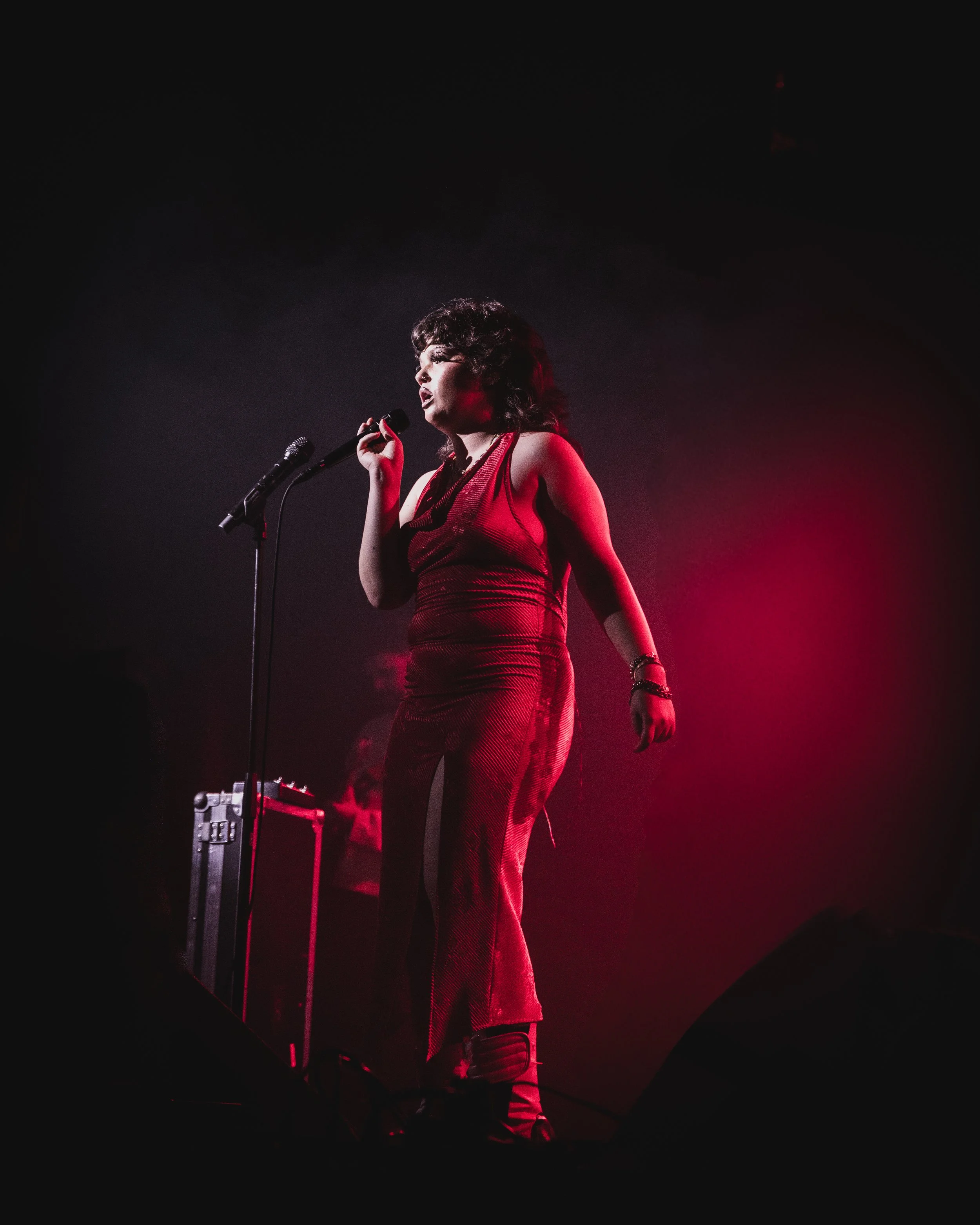 A woman with dark curly hair wearing a red sleeveless outfit singing into a microphone on stage with red lighting.