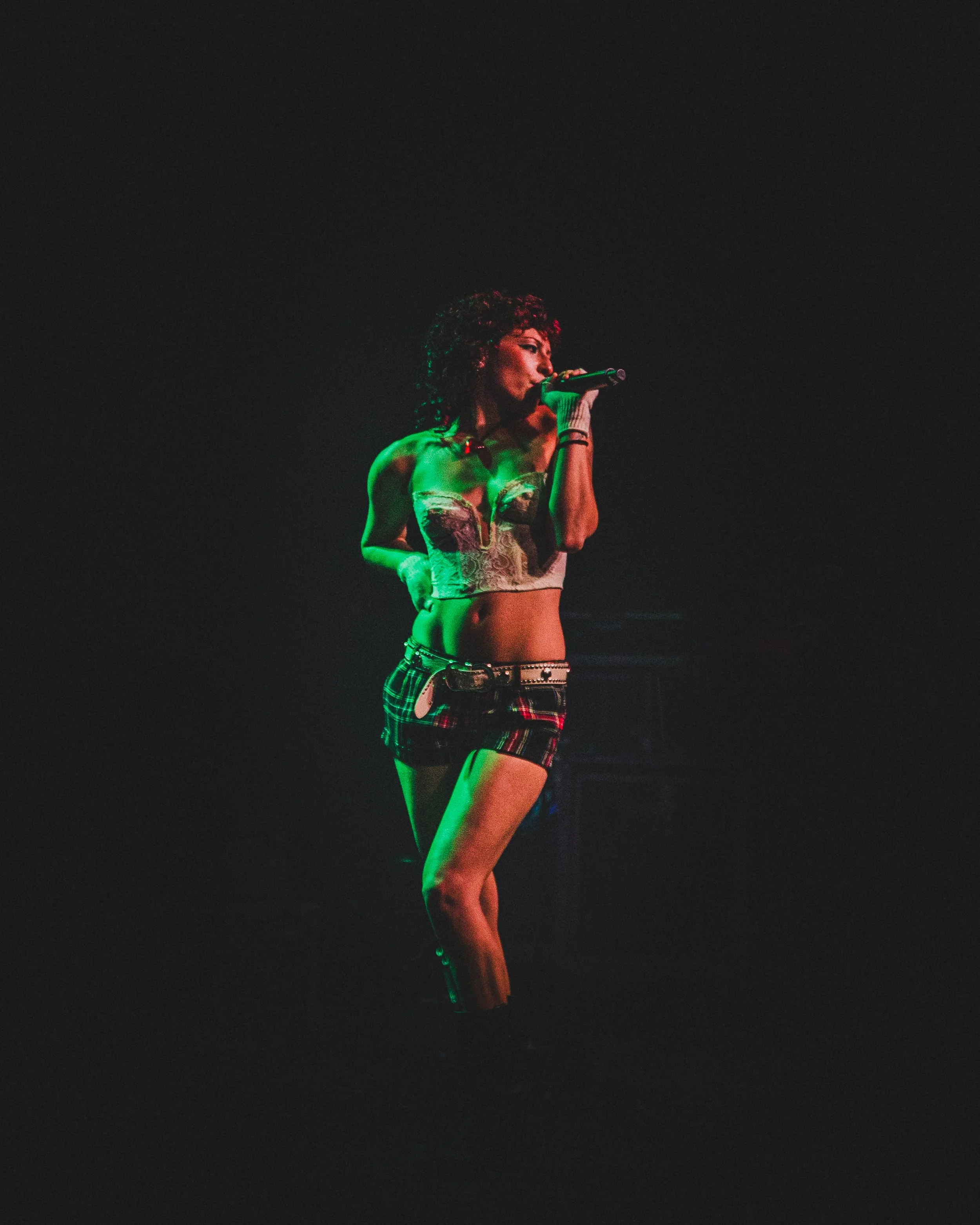 A woman with curly hair singing into a microphone on a dark stage, wearing a bustier top and plaid shorts.