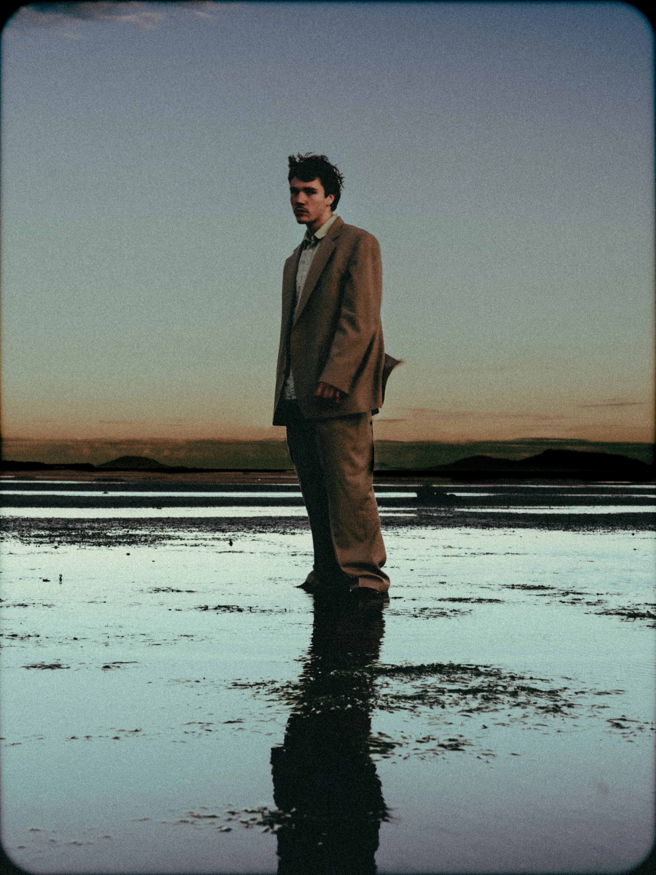 A young man in a brown suit standing on wet ground during sunset with a landscape in the background.