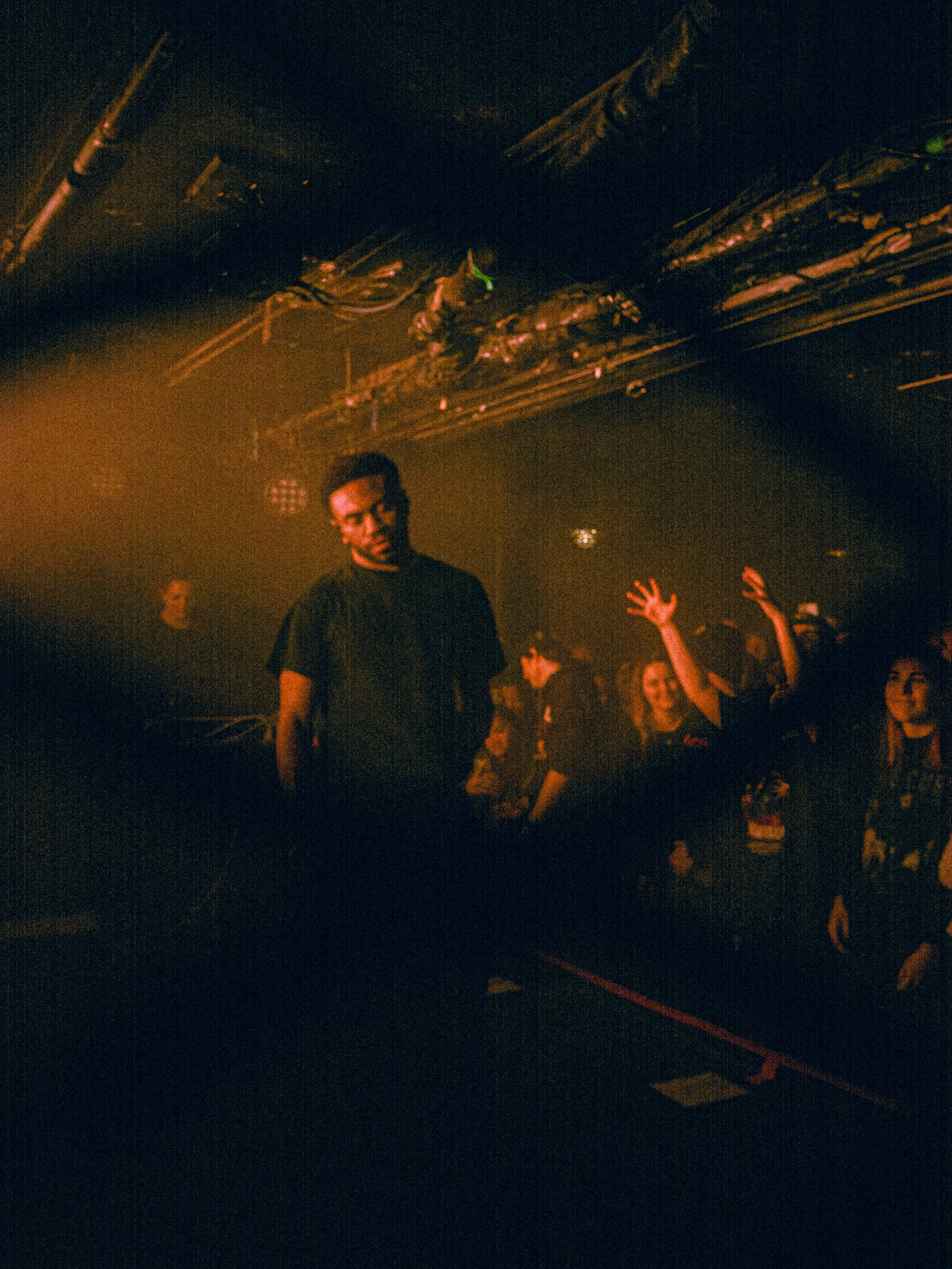 A person standing alone on a dance floor with people dancing and raising their hands, in a dimly lit nightclub or concert venue.