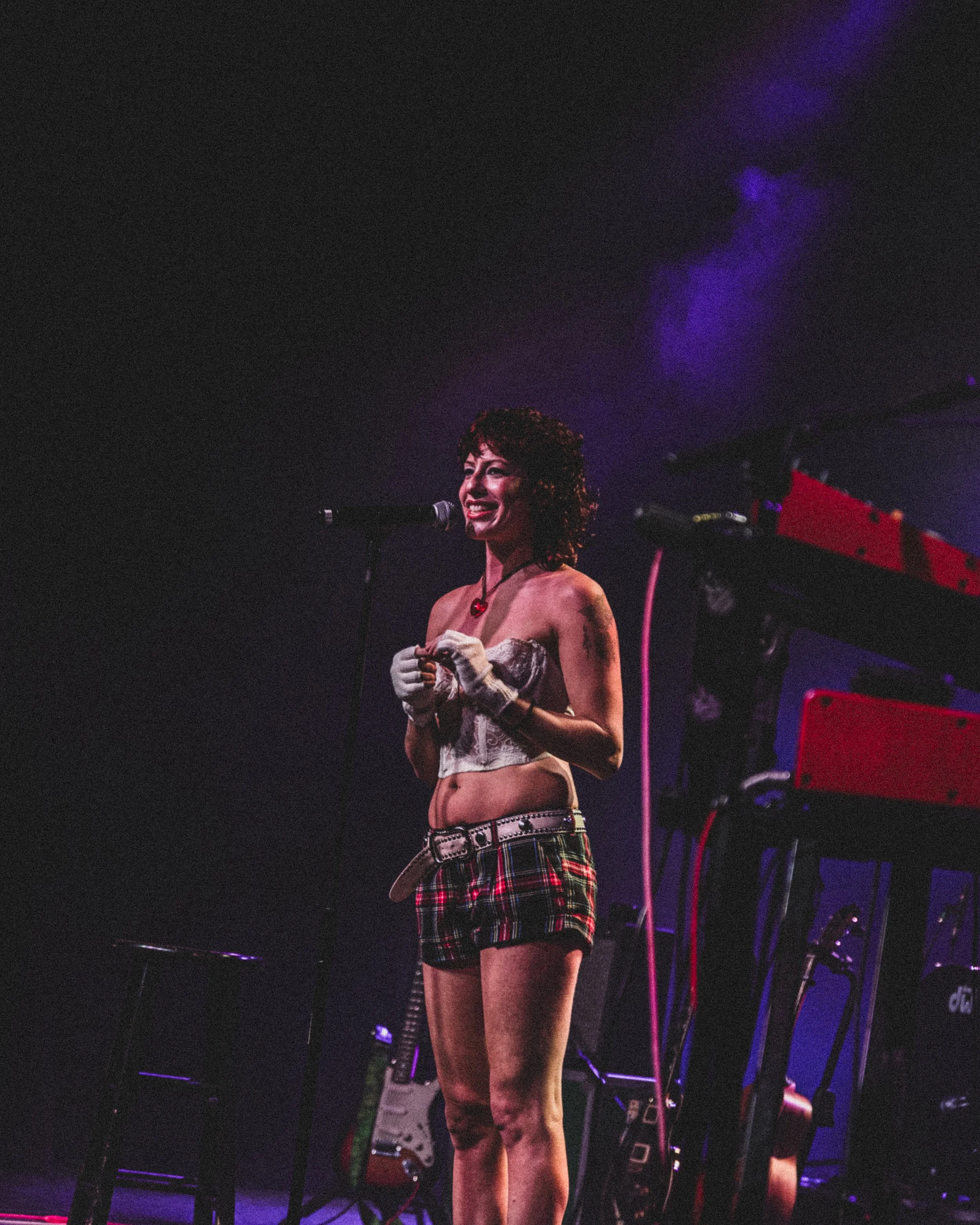 A woman with curly hair smiling and standing on stage in front of a microphone, wearing a lace top, plaid shorts, and gloves, with musical instruments and equipment in the background.