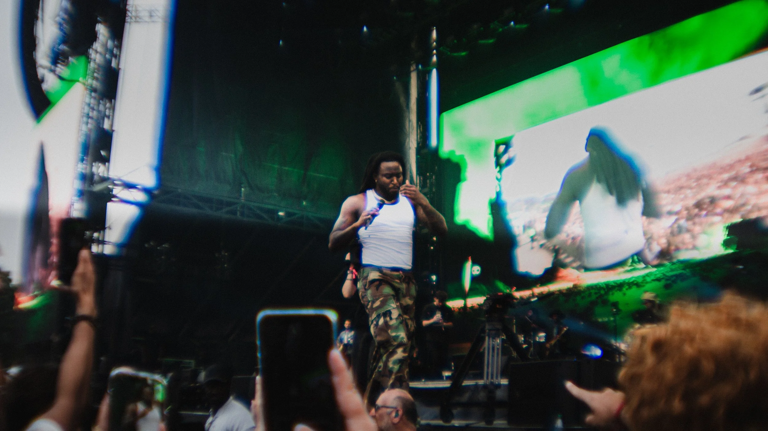 Musician with dreadlocks, wearing a white tank top and camouflage pants, plays a guitar on stage with large screens in the background at an outdoor concert.