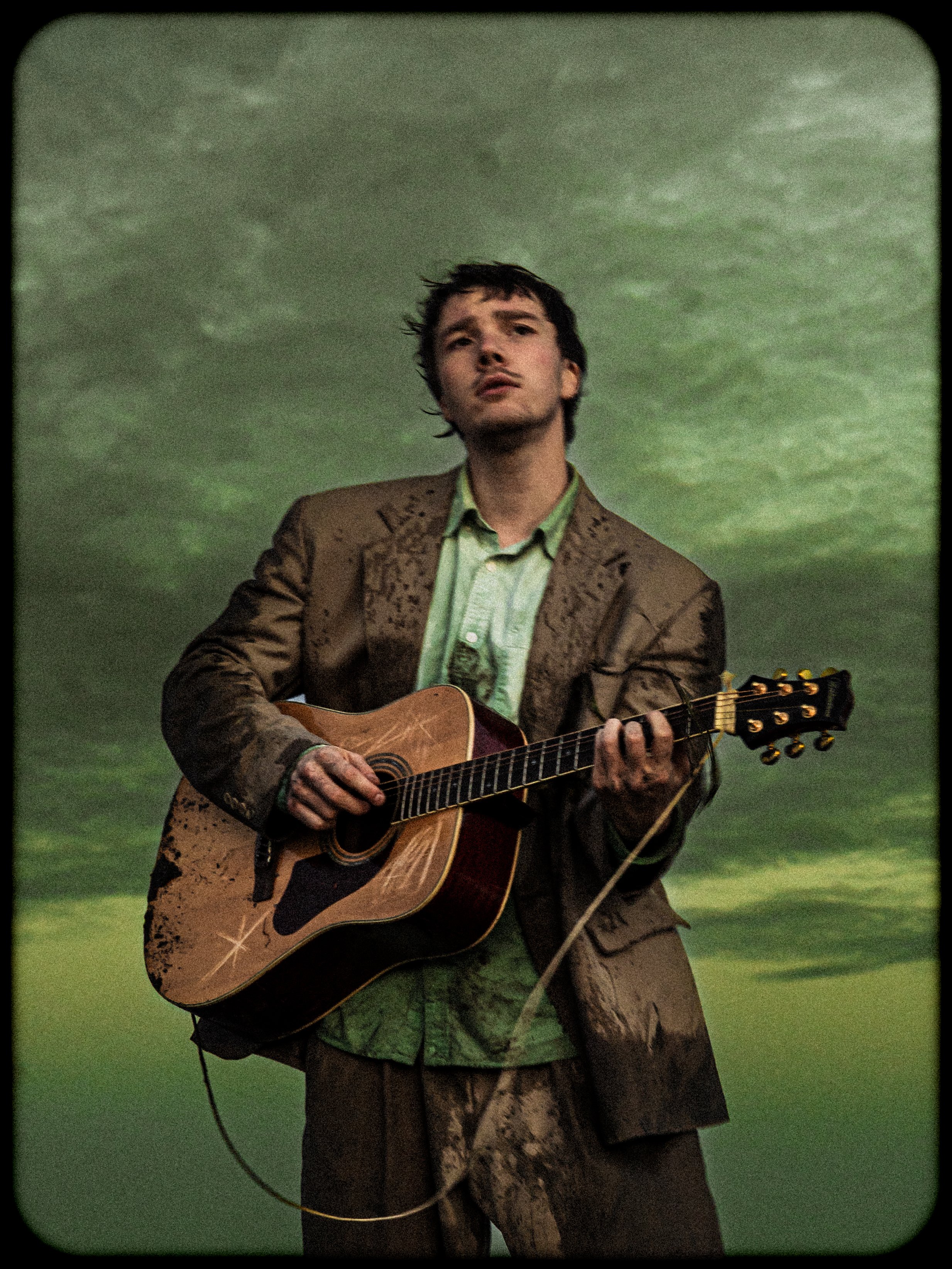 A man playing an acoustic guitar against a cloudy green sky.