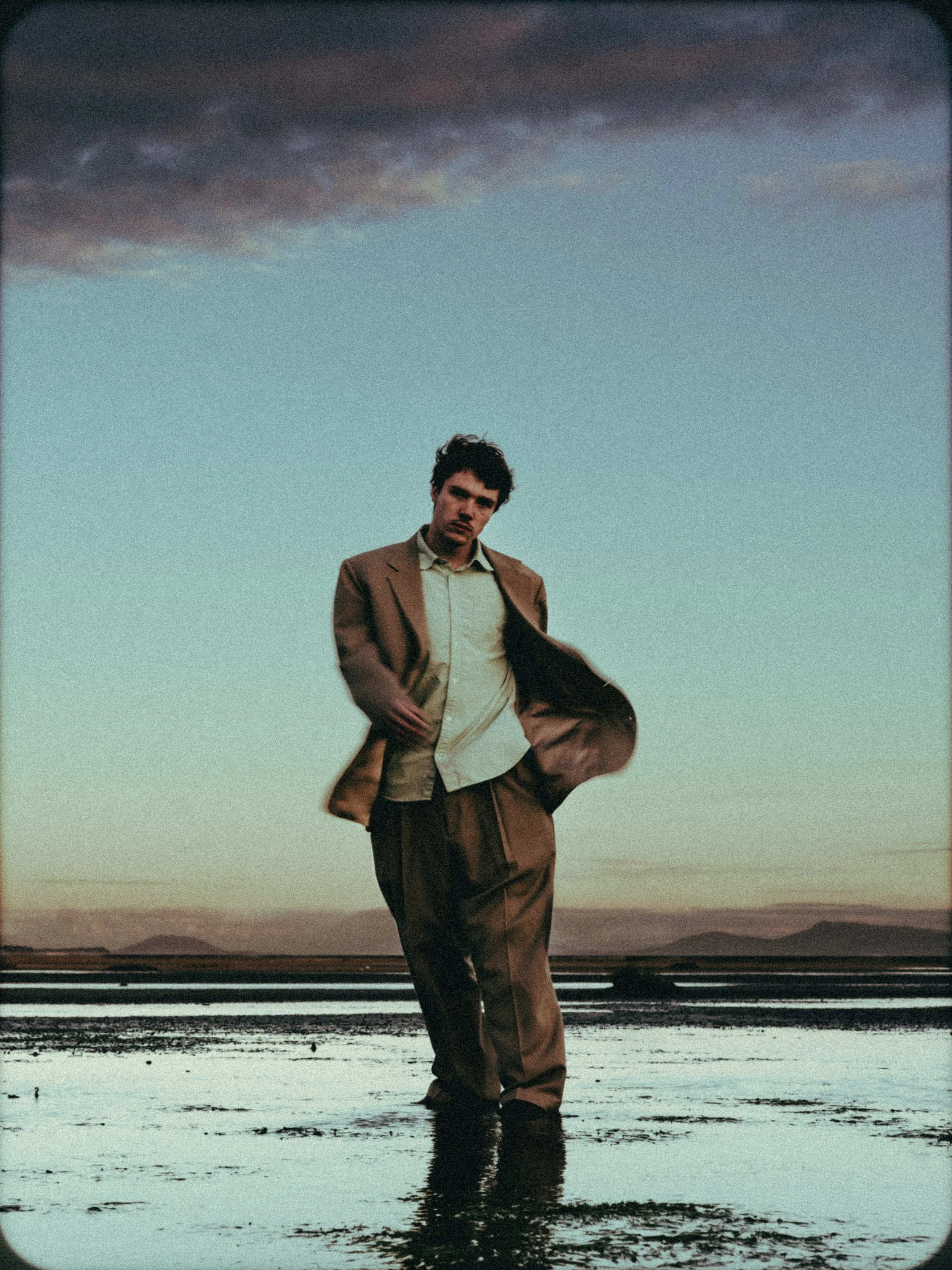 A man in a suit standing in shallow water during sunset with dark clouds overhead.