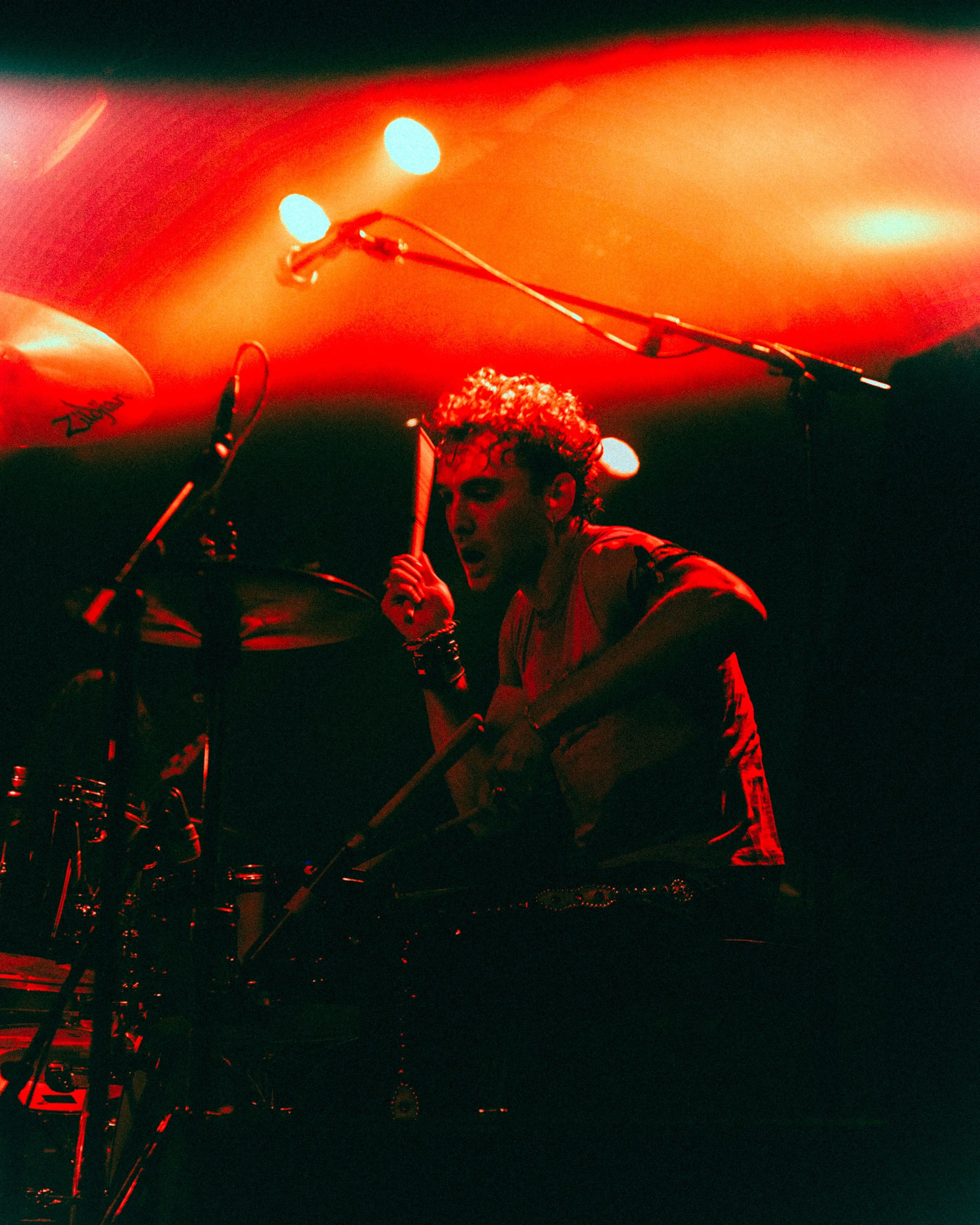 A musician with curly hair passionately playing drums under red stage lighting.