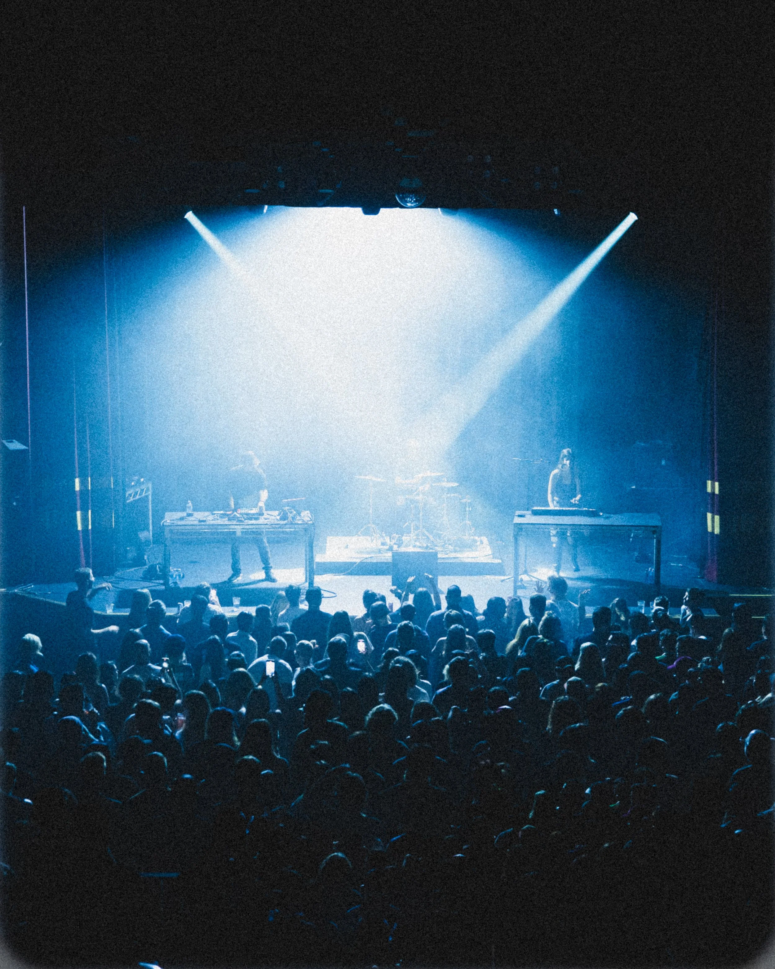 Crowd watching a live electronic music performance on stage with two DJs and drum set, illuminated by blue stage lights