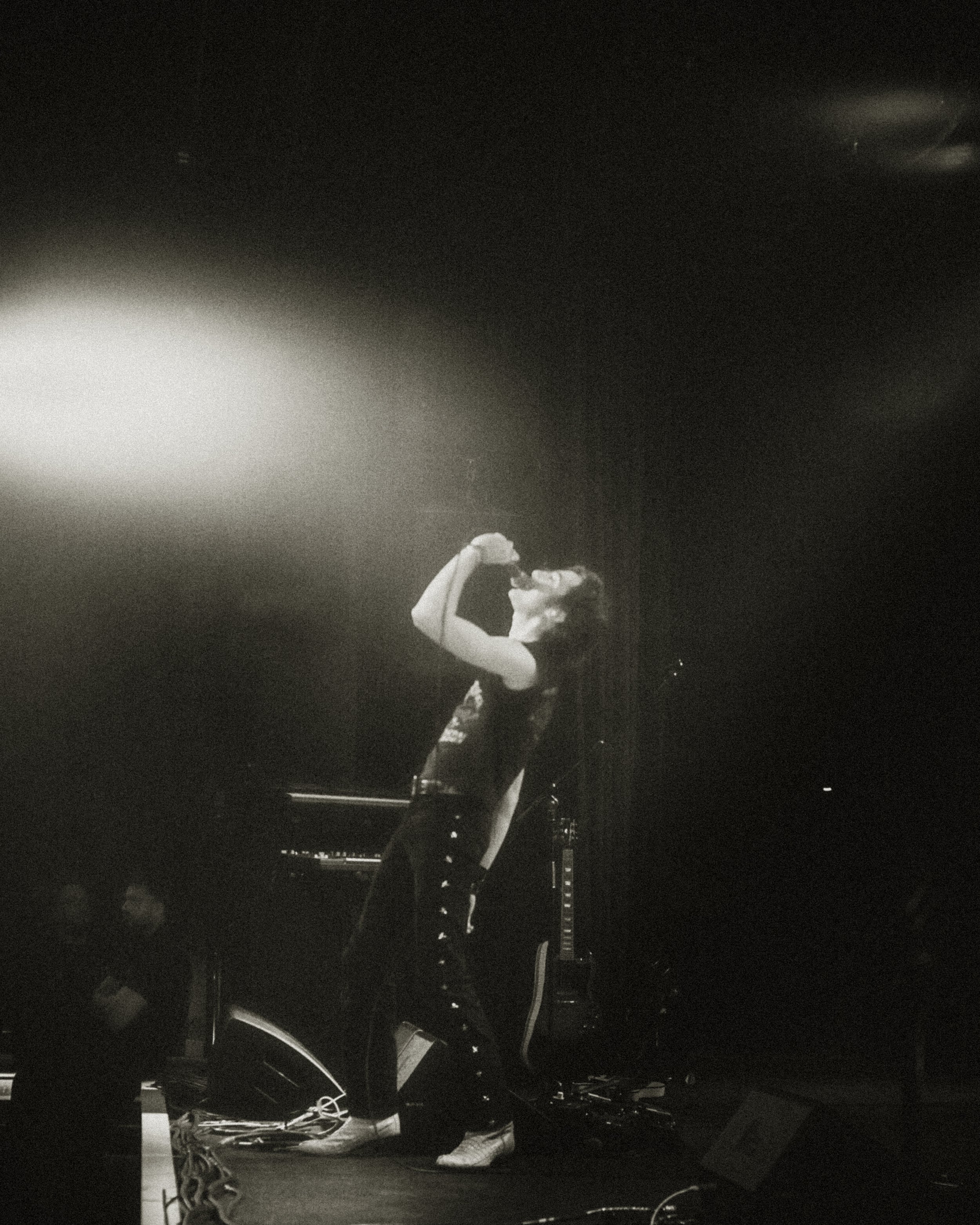 A person on stage singing into a microphone, with musical equipment and guitar in the background, in a dimly lit setting.