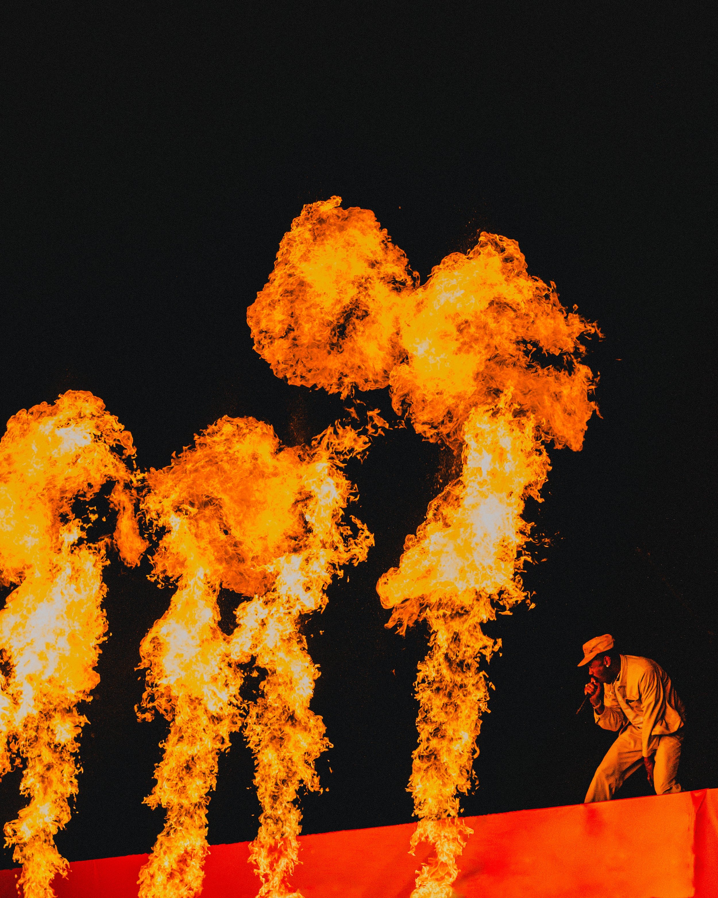 A performer dressed in beige, standing on a platform, appears to be singing or speaking into a microphone amid large flames against a dark night sky.