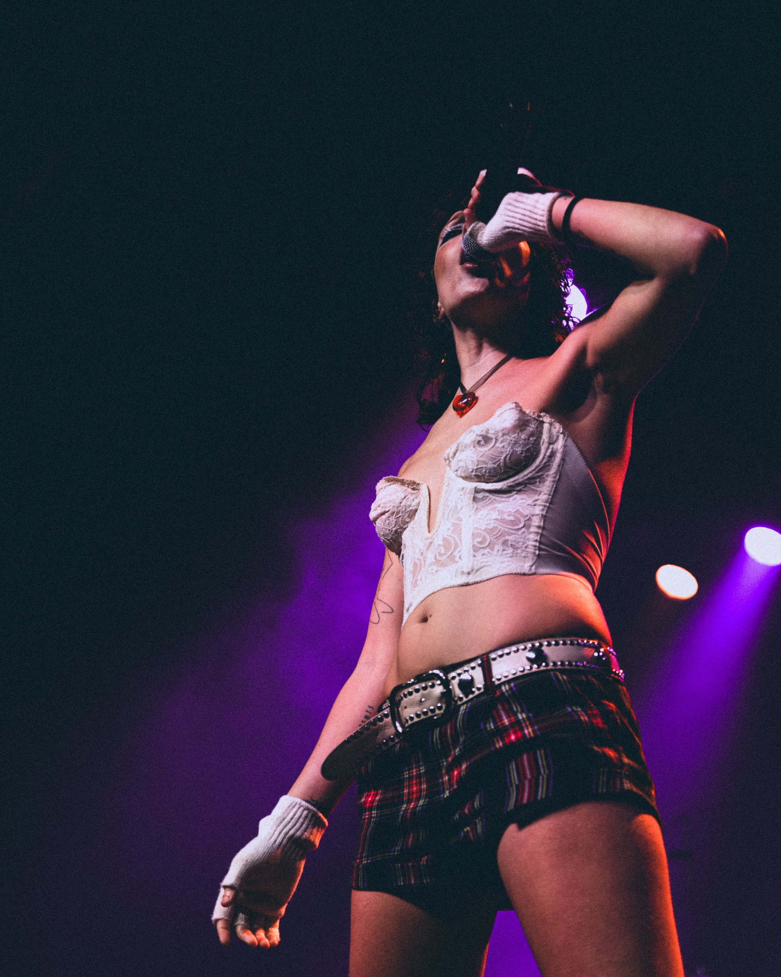 Performer singing on stage with purple lighting, wearing a white lace bustier top, plaid shorts, and fingerless gloves.