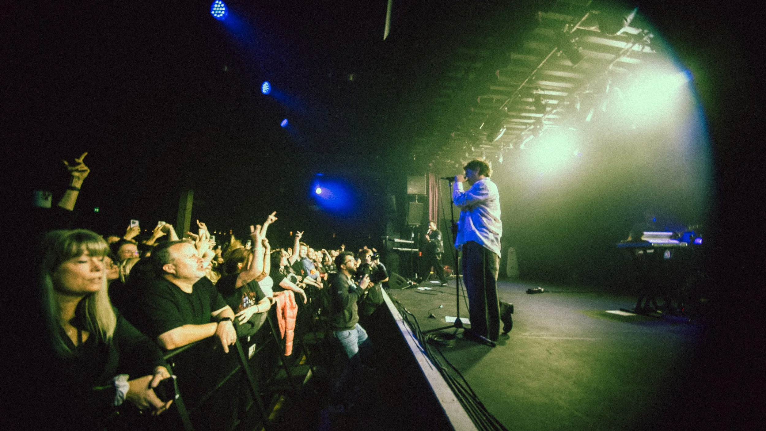 A performer singing on stage with audience members enjoying and some raising their hands during a concert with stage lights.