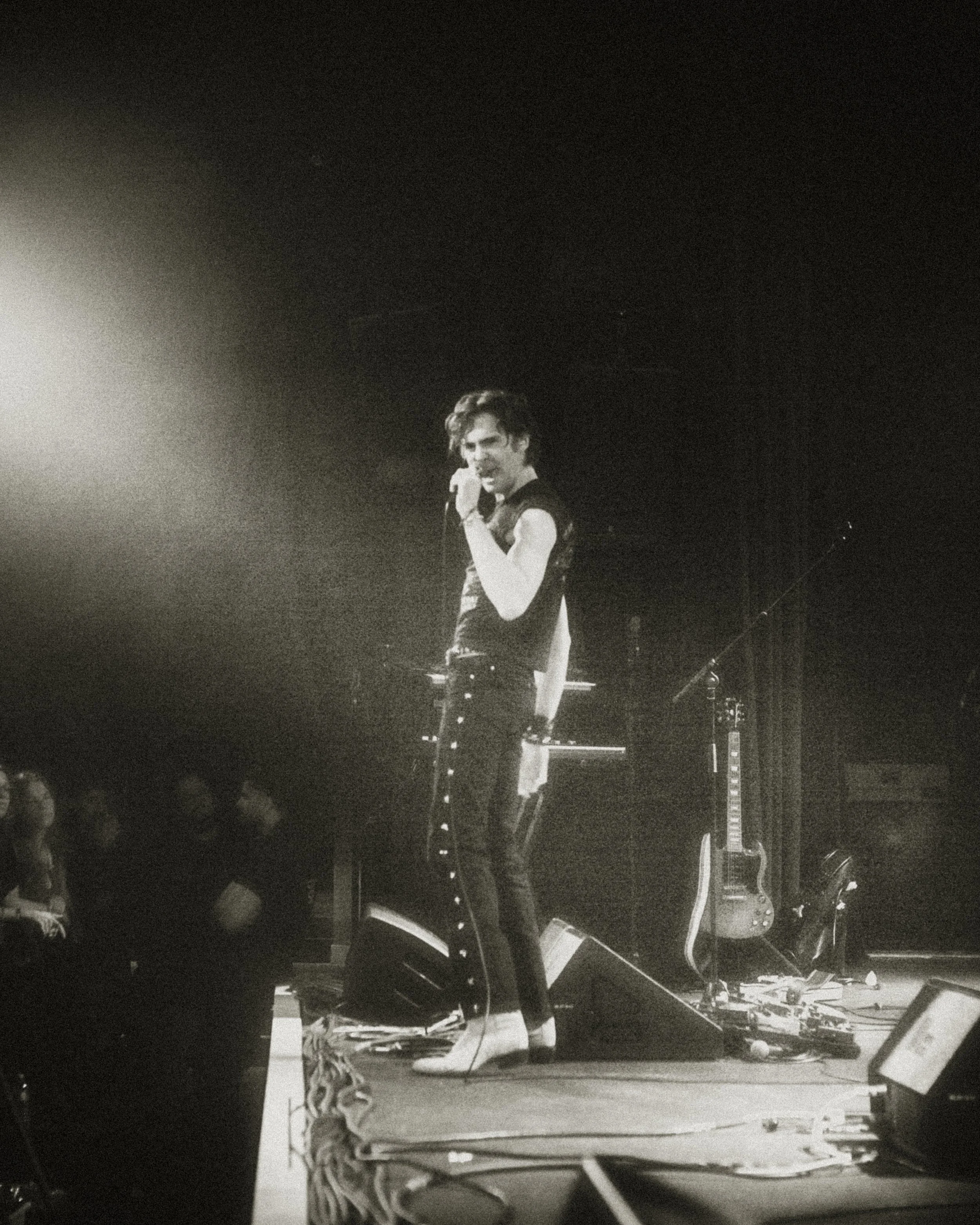 A musician performs on stage with a microphone, standing next to musical instruments, including guitars. The scene is dimly lit with a spotlight.