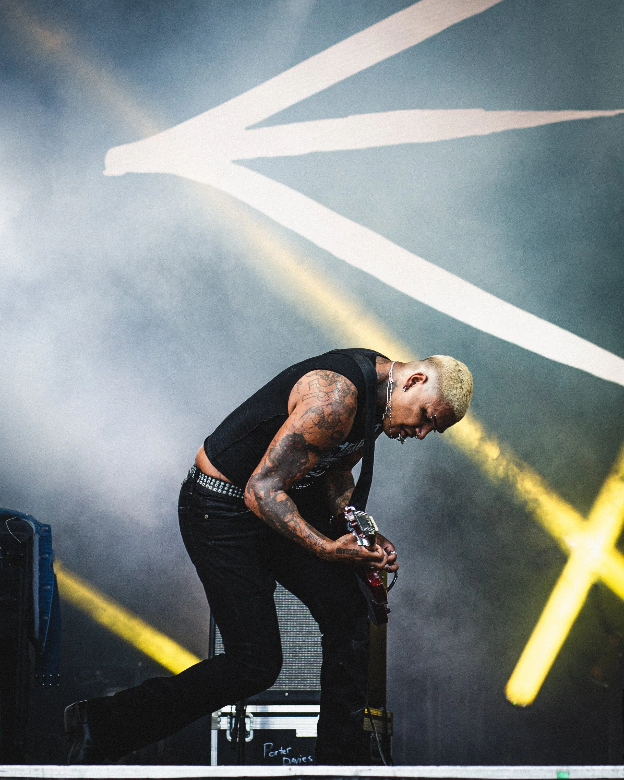 A musician with bleached blonde hair and tattoos on his arms playing an electric guitar on stage, with yellow lights and a dark background.