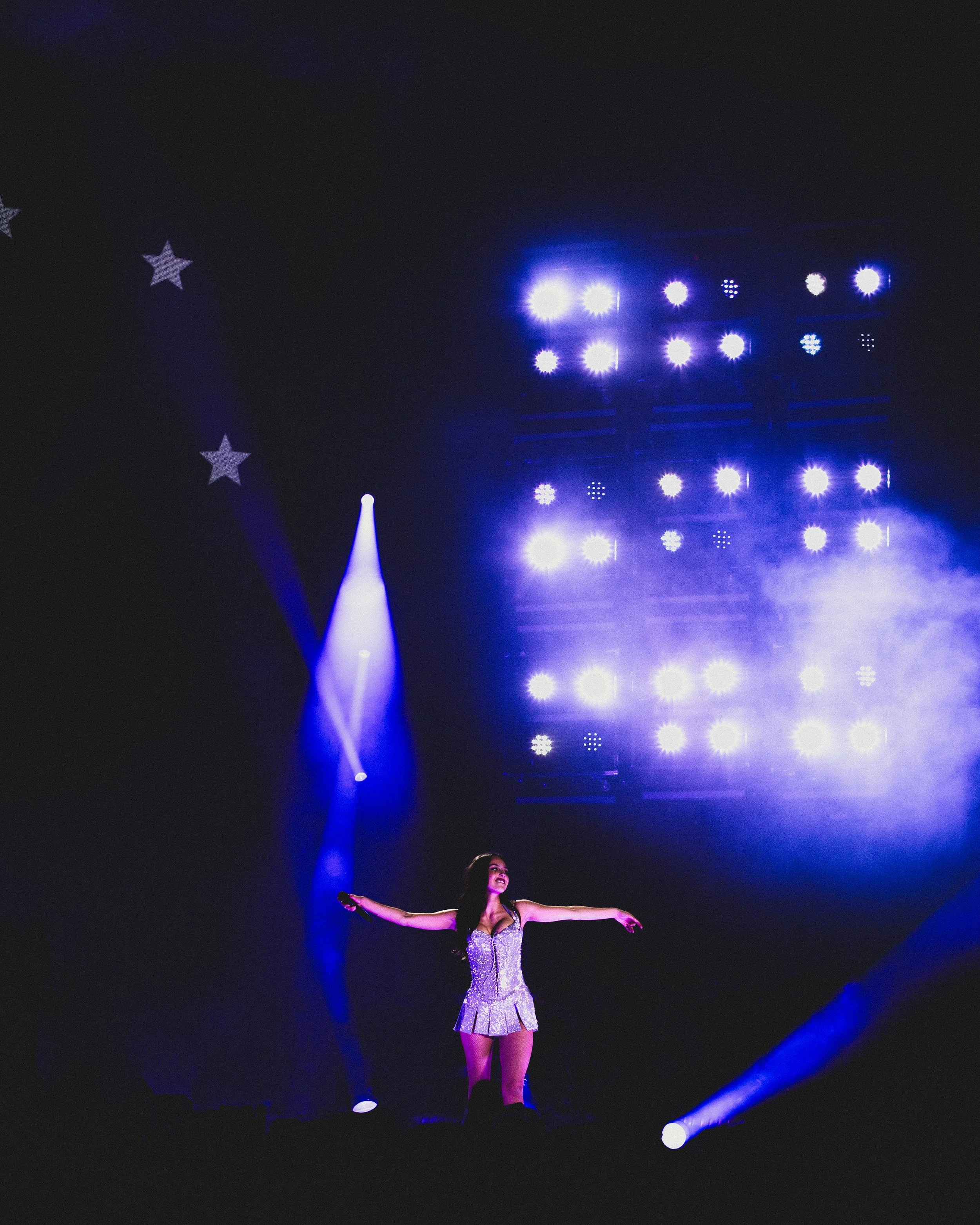 A woman performing on stage with her arms outstretched, illuminated by bright stage lights, with a dark background that includes some stars.