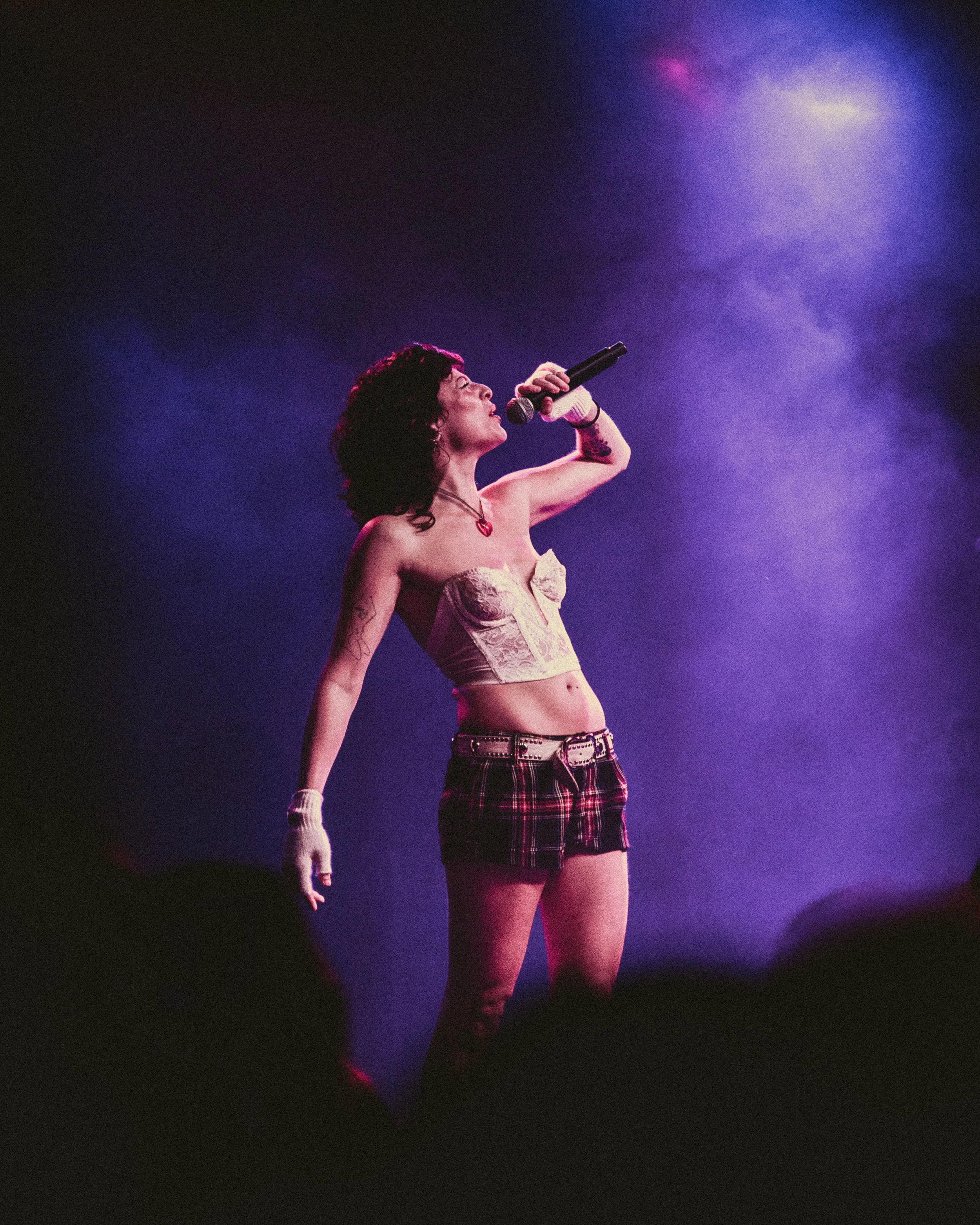 A woman with curly hair singing into a microphone on stage, wearing a strapless top, plaid shorts, and a belt.