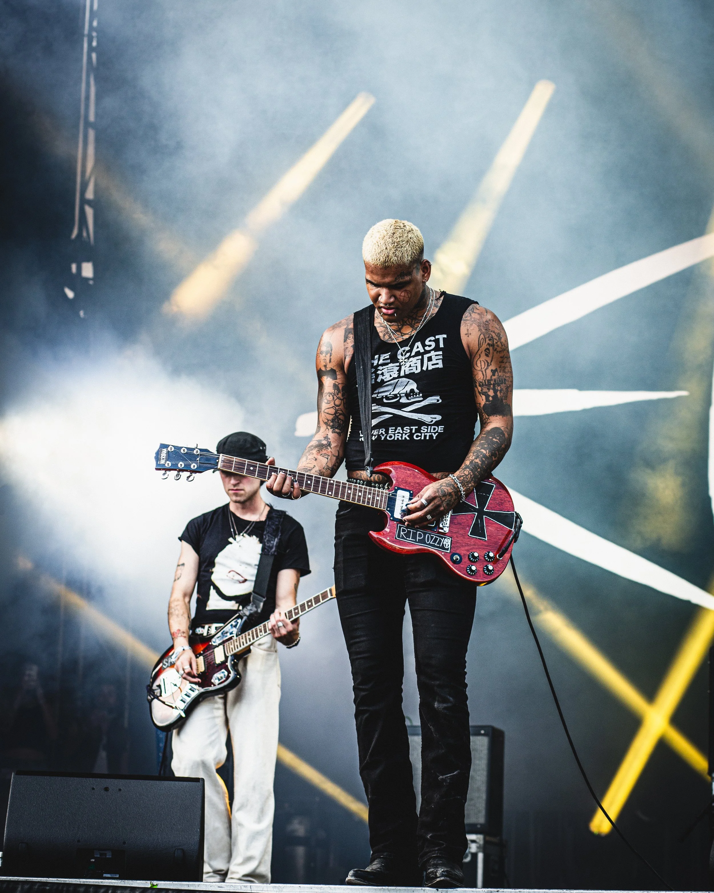 Two musicians performing on stage with guitars, one in the foreground with tied-dye pants, tank top, and tattoos, and another with a black T-shirt and light-colored pants, both under stage lights and smoke.