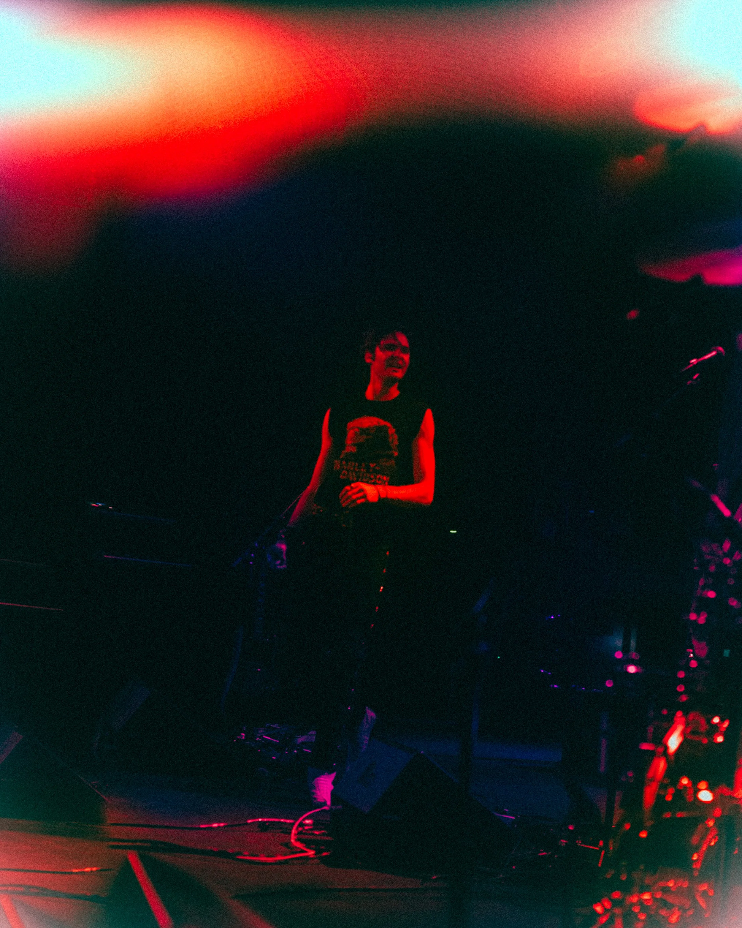 A person standing on a dark stage with red and blue lights, wearing a black sleeveless shirt with a graphic, holding a microphone, and surrounded by musical equipment.