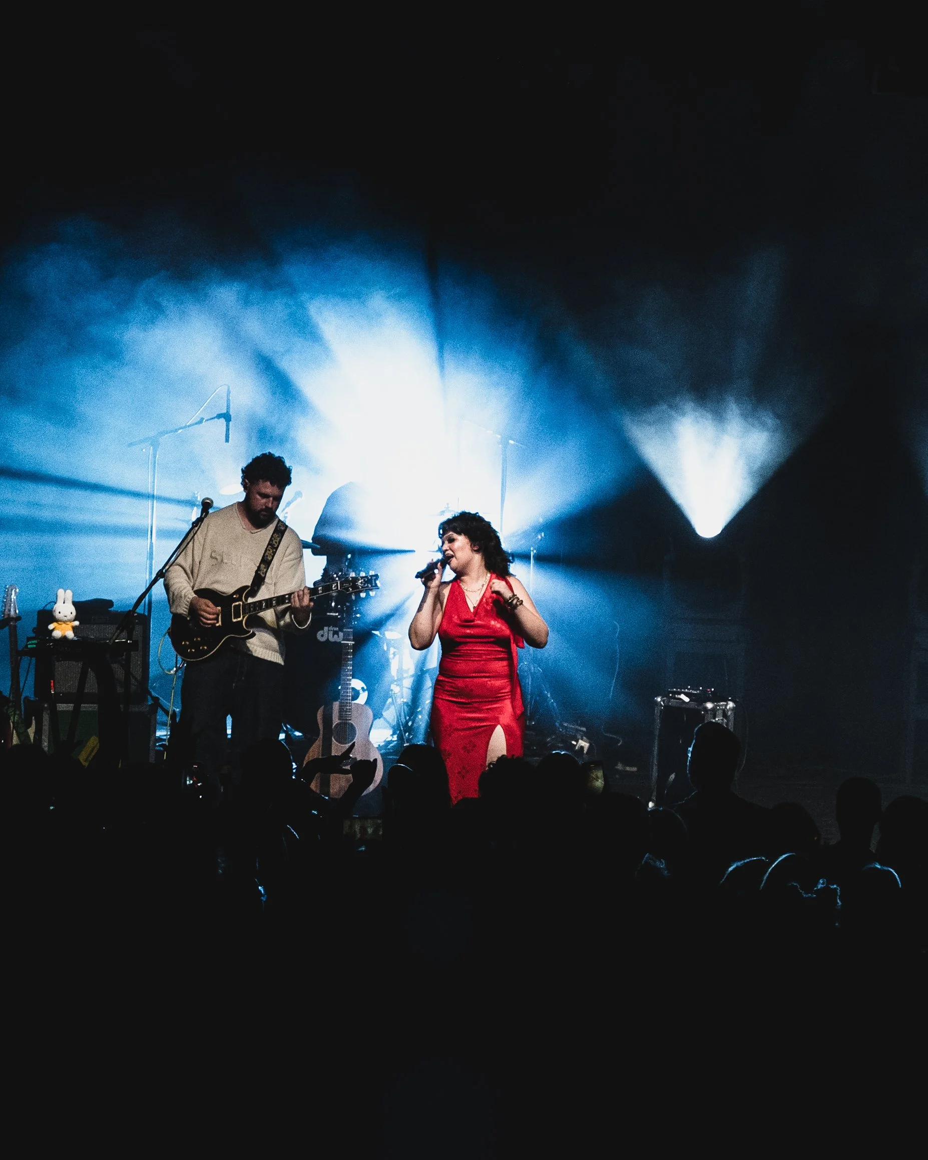 A live music performance with a singer in a red dress and a guitarist on stage, with blue lighting and an audience in front.
