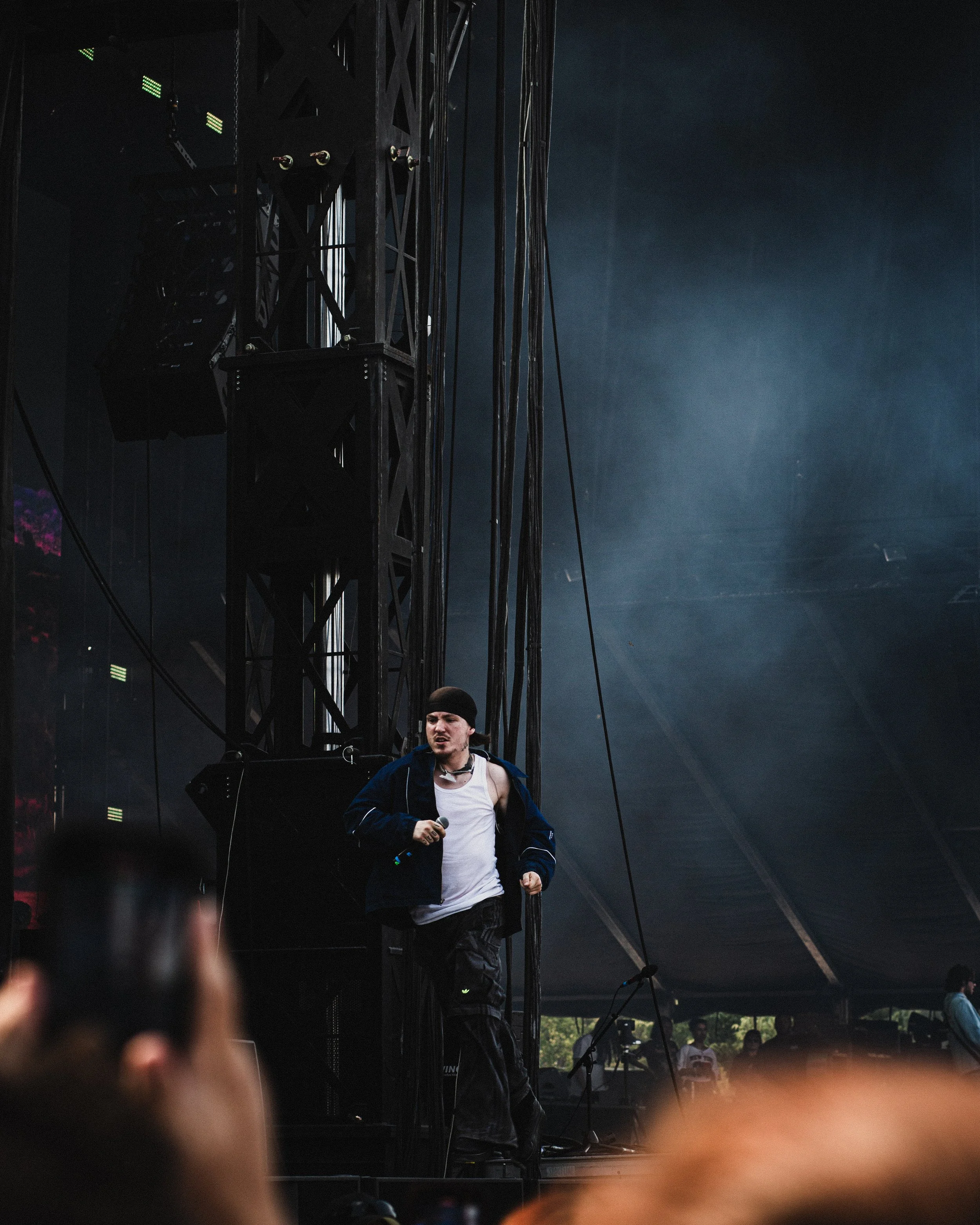 A performer holding a microphone standing on stage at a music concert under a large black canopy, with stage equipment and scaffolding visible.