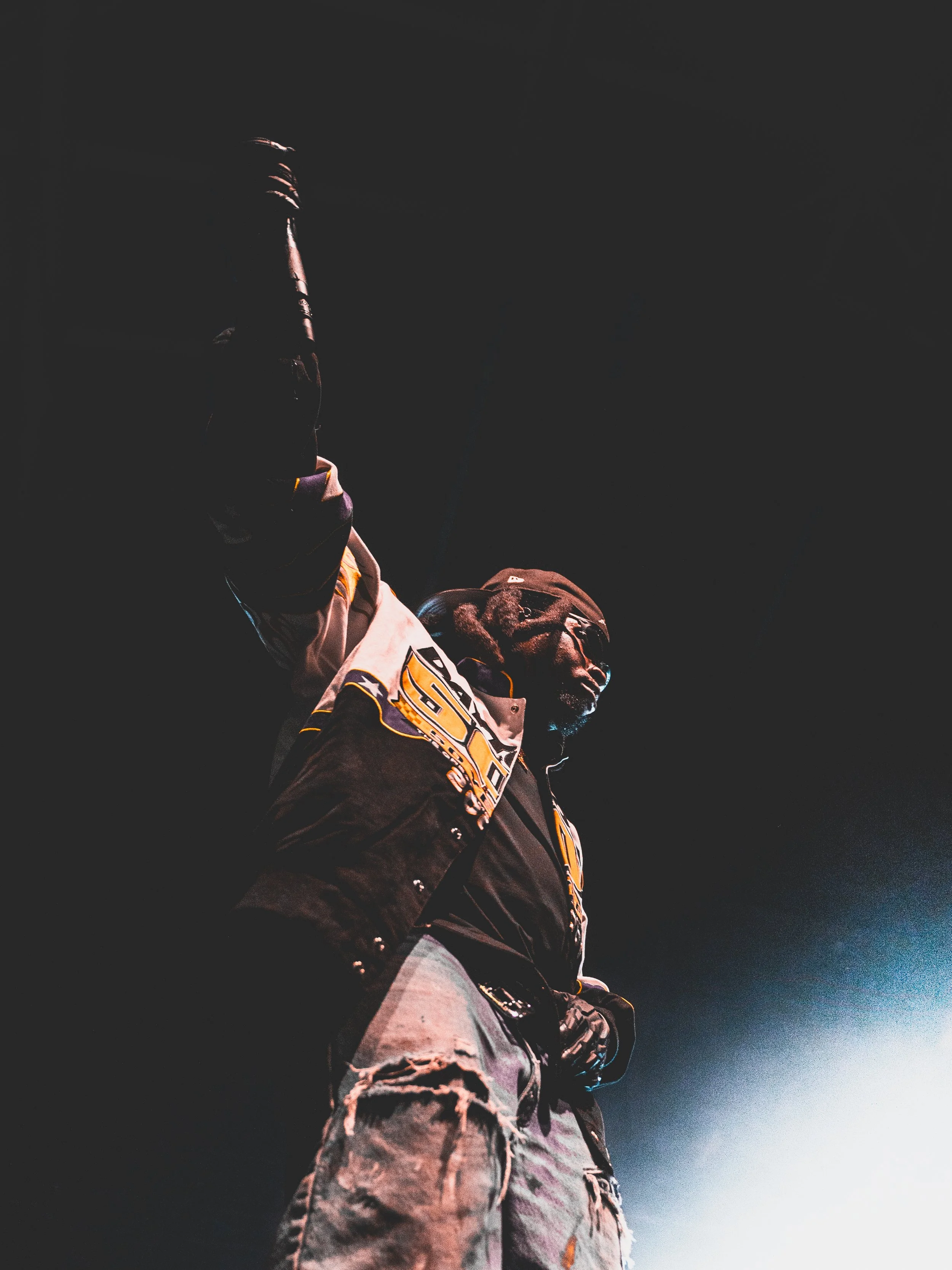 A person dressed in a black and white varsity jacket with patches and ripped jeans, wearing sunglasses and a cap, standing against a dark background with a light source illuminating their lower body.