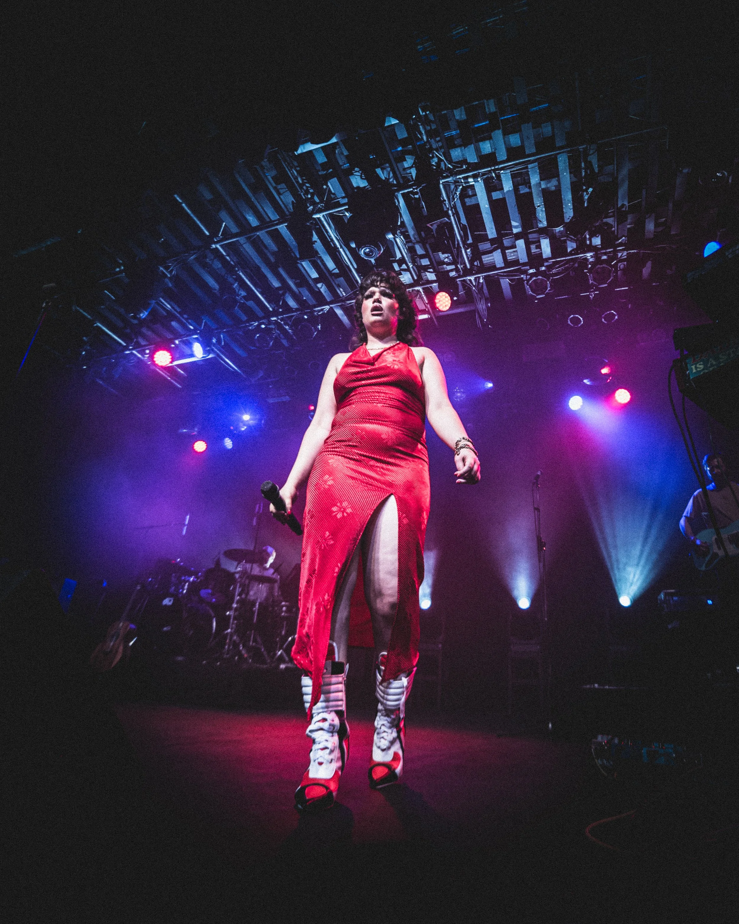 Performer in a red dress and platform boots holding a microphone on a stage with colorful lighting and a band in the background.