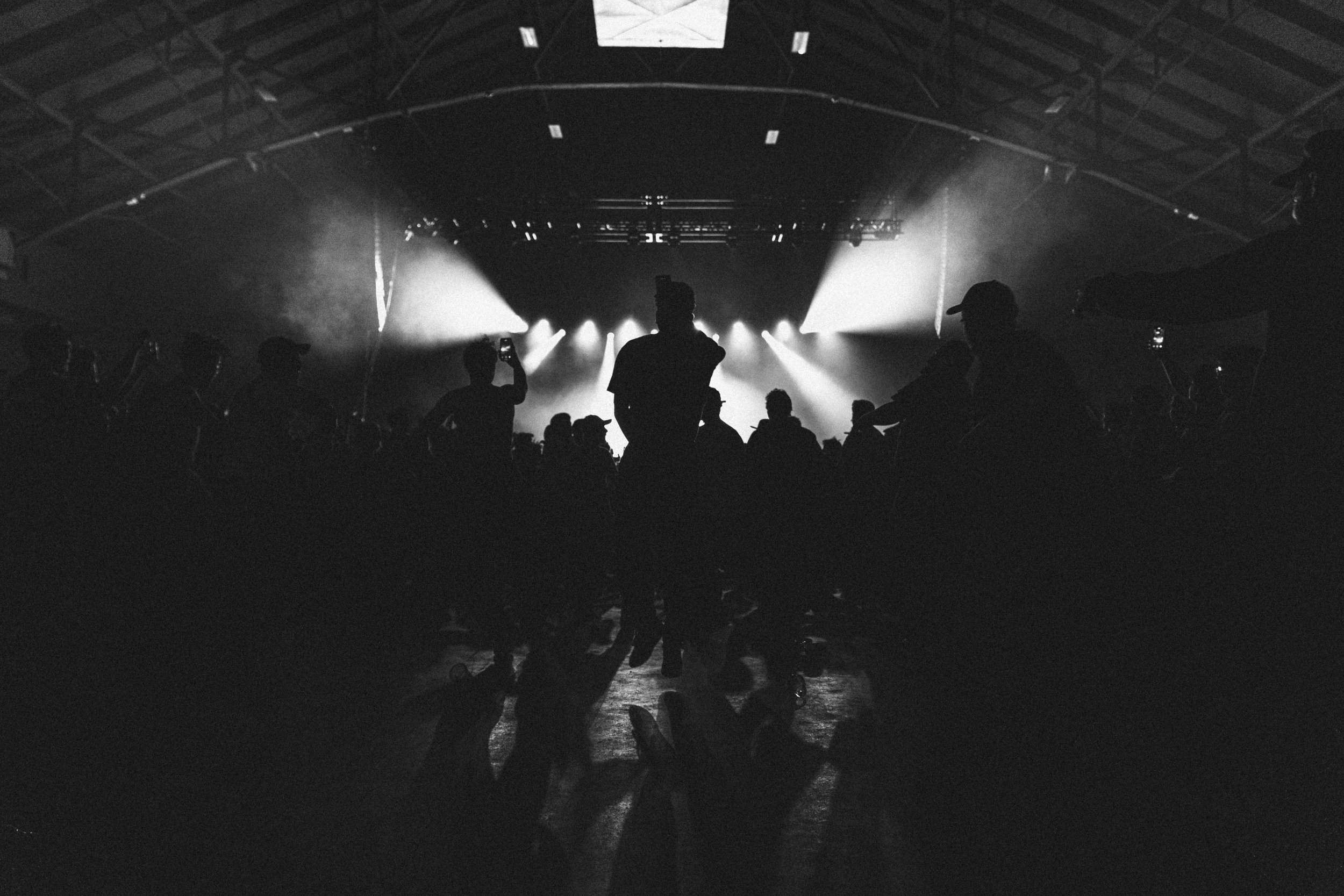 Silhouette of people at a concert or event with stage lights shining in the background, in a dark indoor venue.