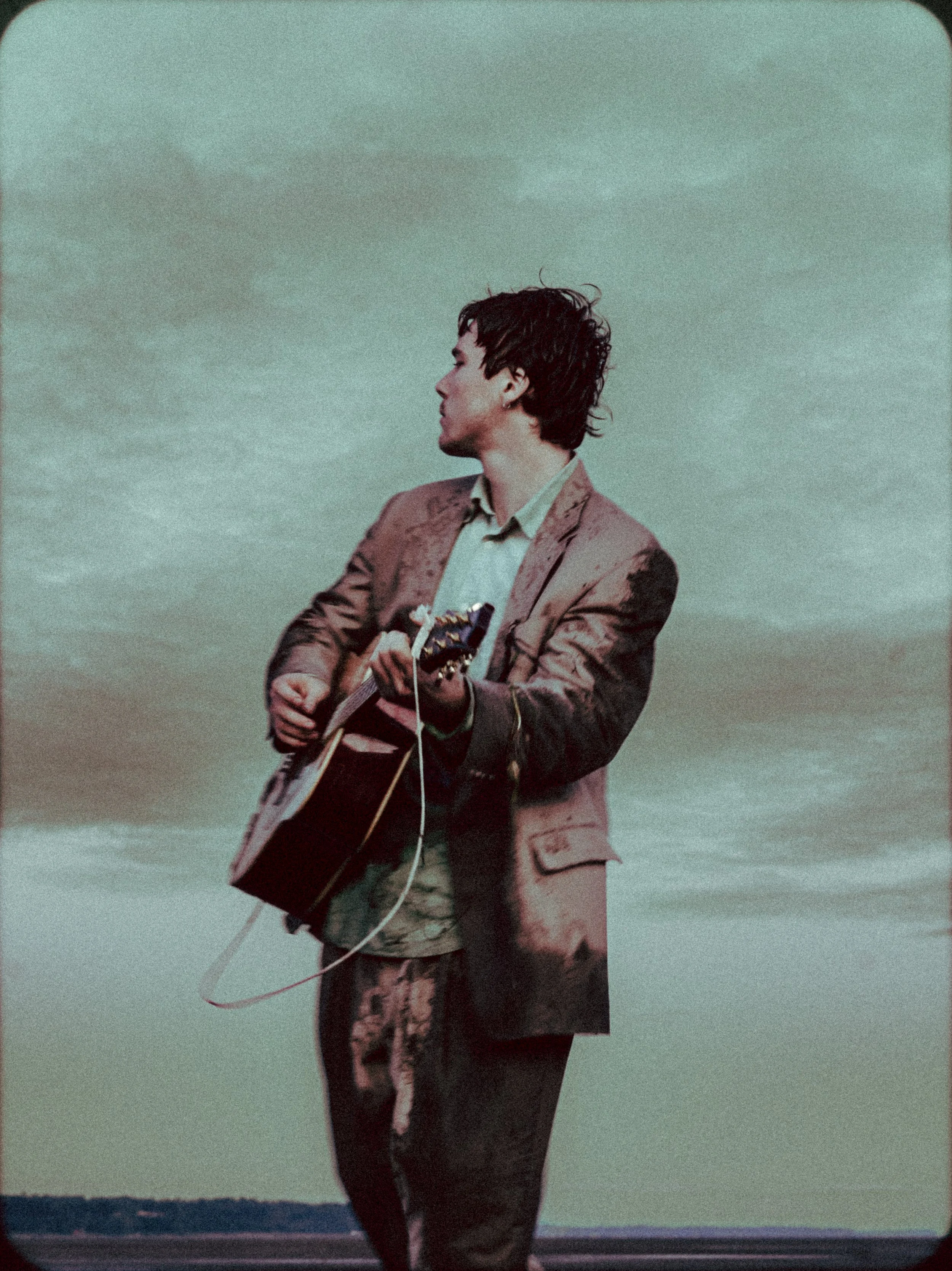 A man in a brown jacket playing an acoustic guitar outdoors against a cloudy sky.