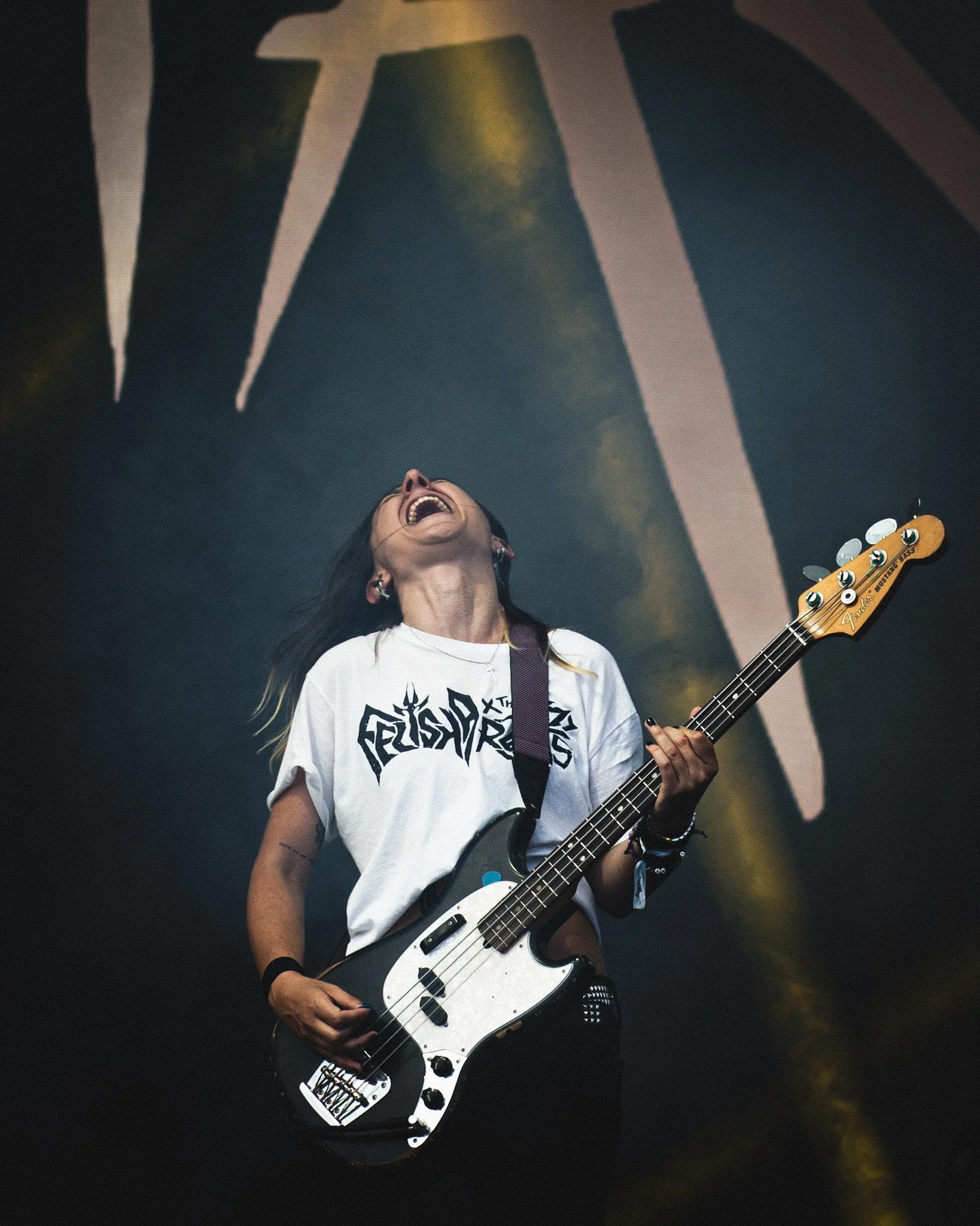 Female musician playing a bass guitar on stage, wearing a white t-shirt with band logo, laughing or singing, with a large stage light and logo in the background.
