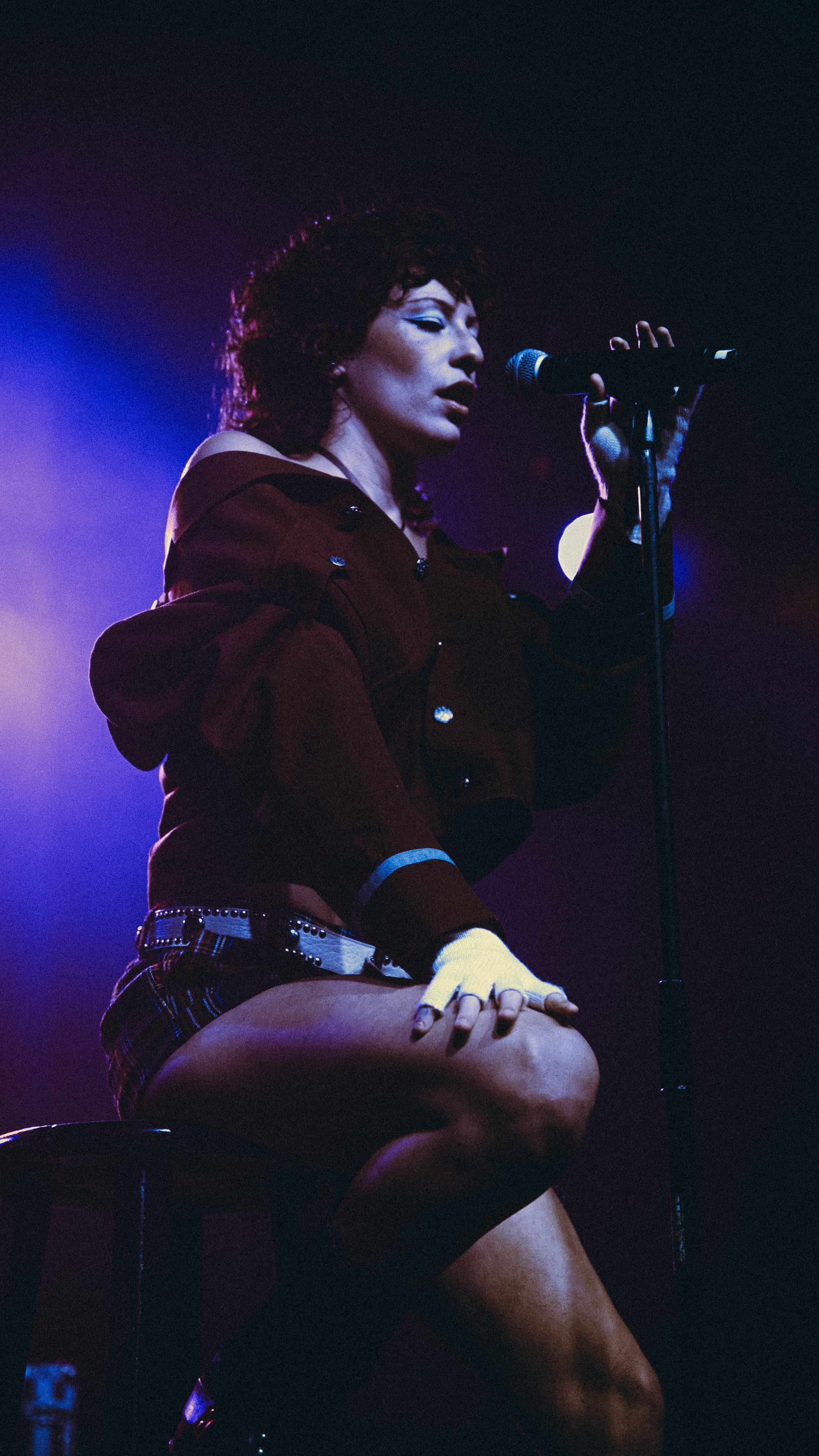 A woman performing on stage, sitting on a stool, singing into a microphone, wearing a red jacket, plaid shorts, and a glove.