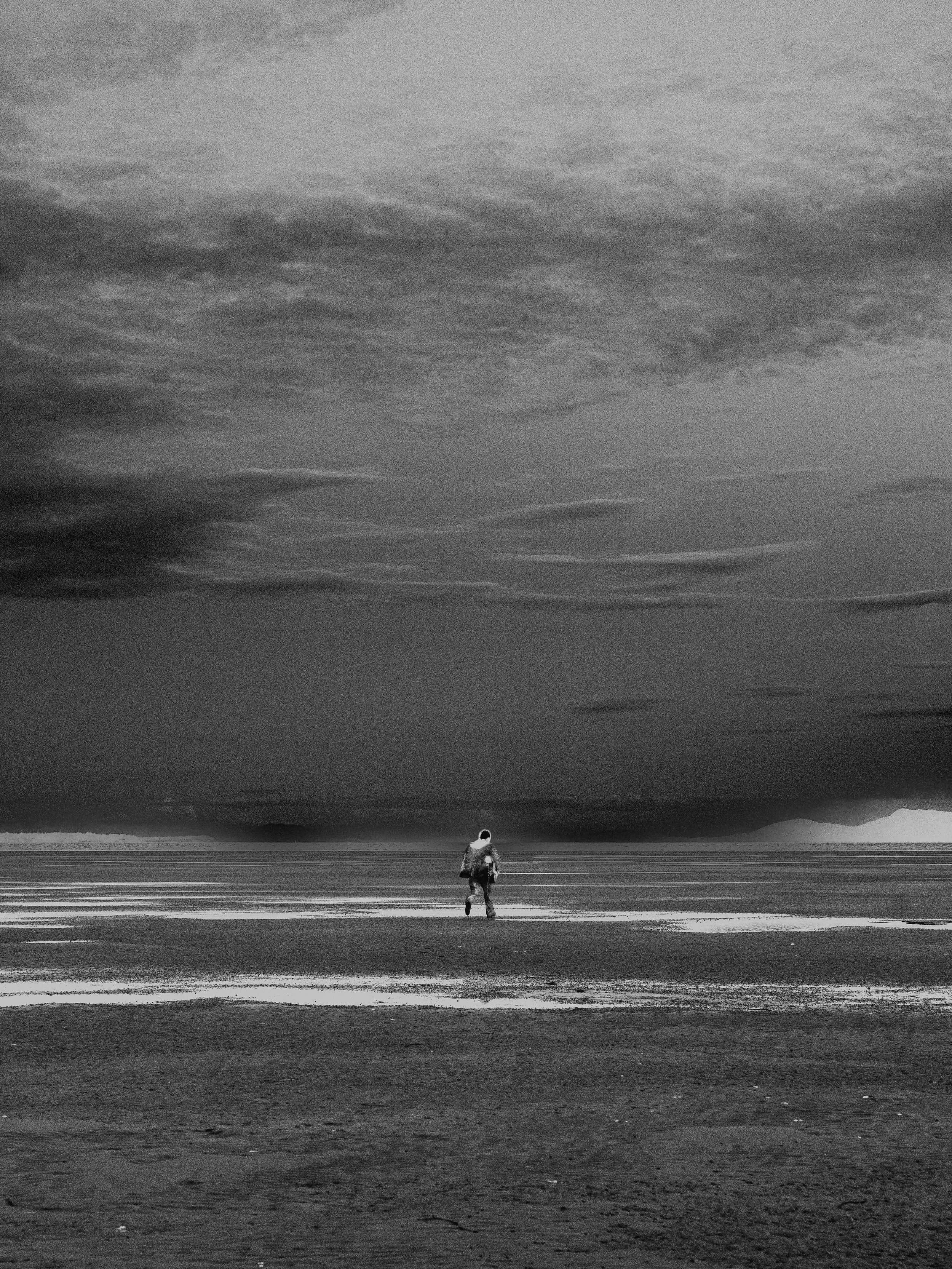 A person walking alone on a beach near the water, with dark clouds in the sky, in black and white.